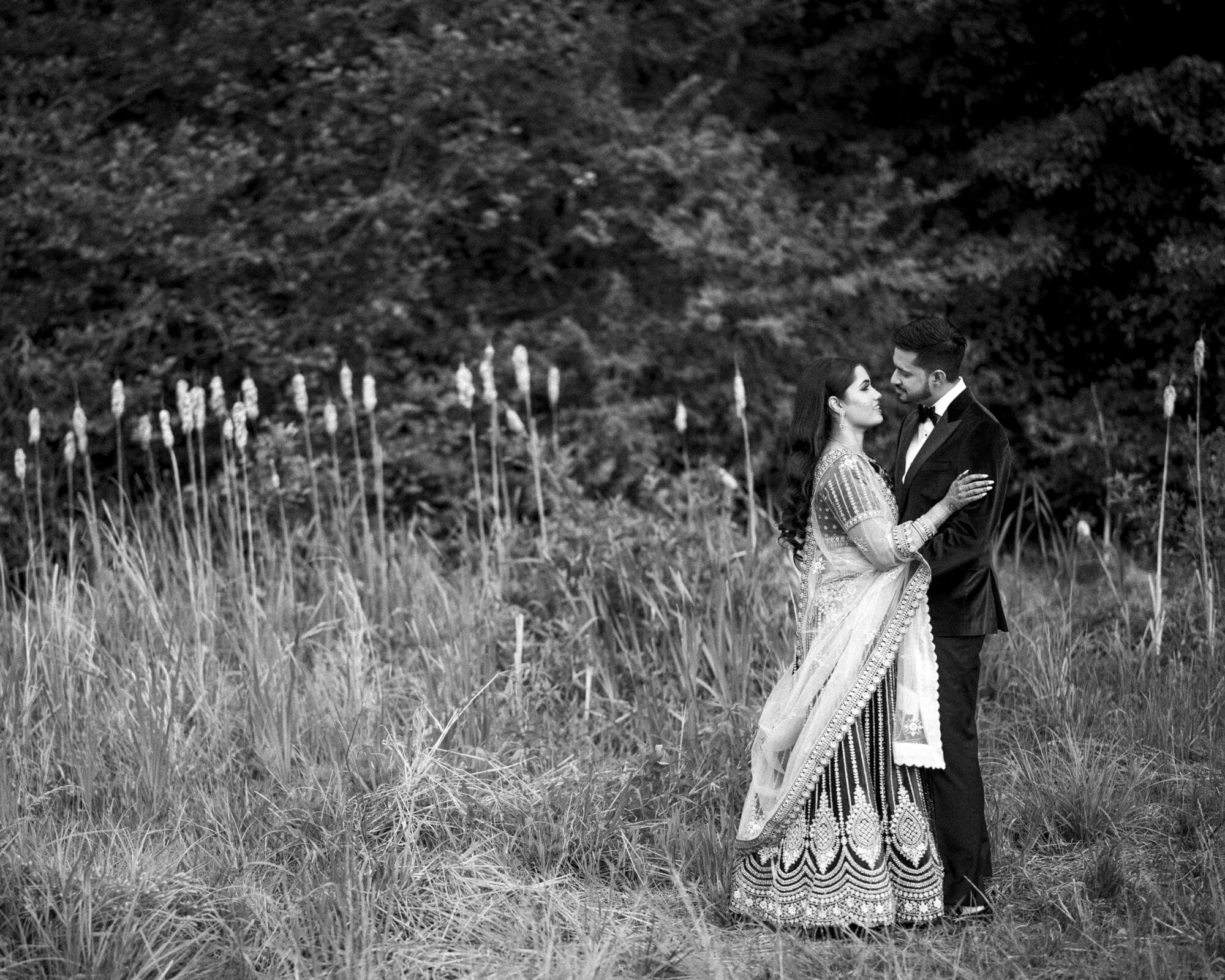 A romantic black and white wedding portrait of a couple embracing among tall ornamental grasses on a beautiful estate.