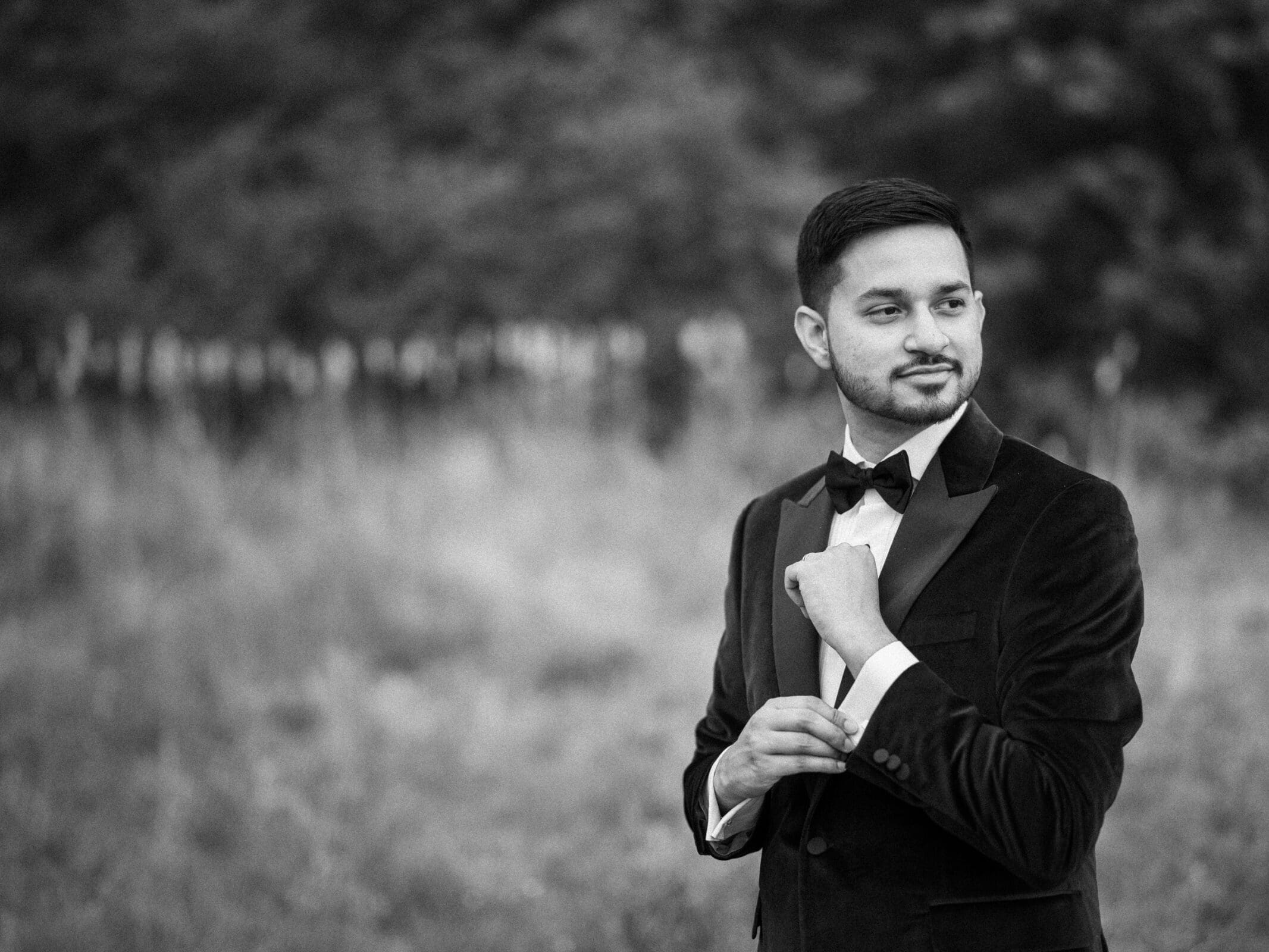 A dapper black and white portrait of a groom adjusting his tuxedo cufflink outdoors before his wedding reception.