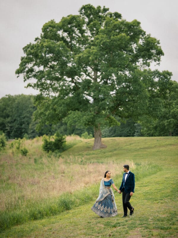 A romantic wide shot of a bride and groom holding hands and walking across a vast, open grassy field on the grounds of Donovan Manor.