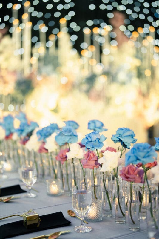 A luxury wedding reception table setting featuring rows of vibrant blue and pink roses under twinkling string lights at Donovan Manor.