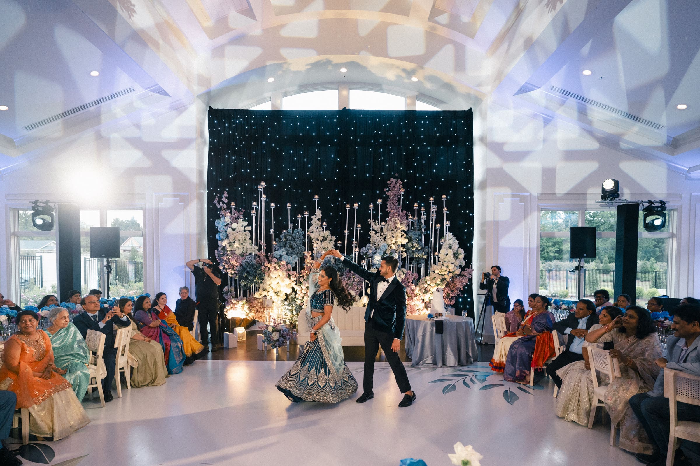 A bride and groom in elegant Indian attire perform a choreographed dance on a decorated ballroom stage during their reception at Donovan Manor.