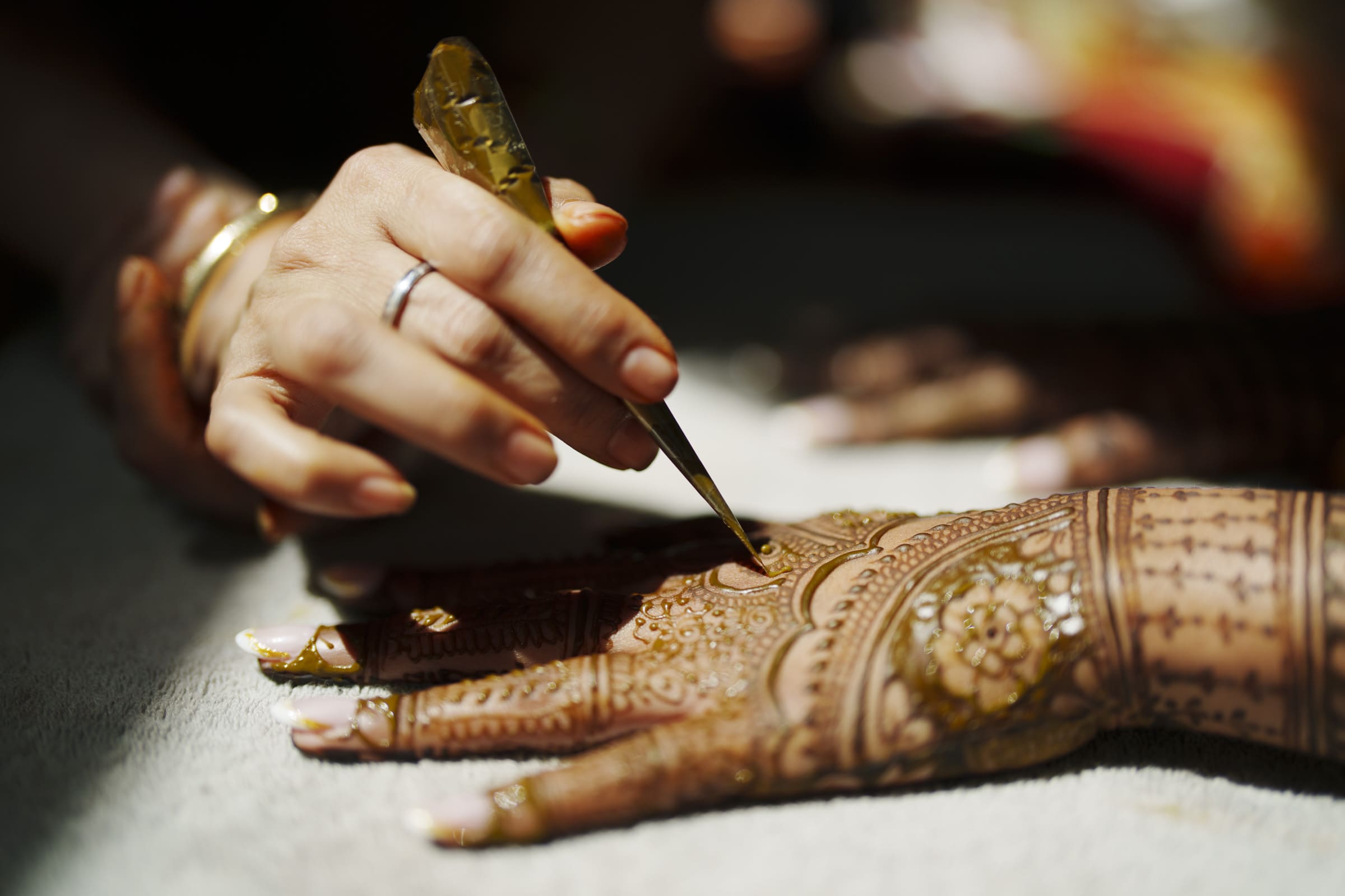 A close-up of a person applying intricate henna designs on another persons hand using a cone, with detailed patterns visible on the palm and fingers.