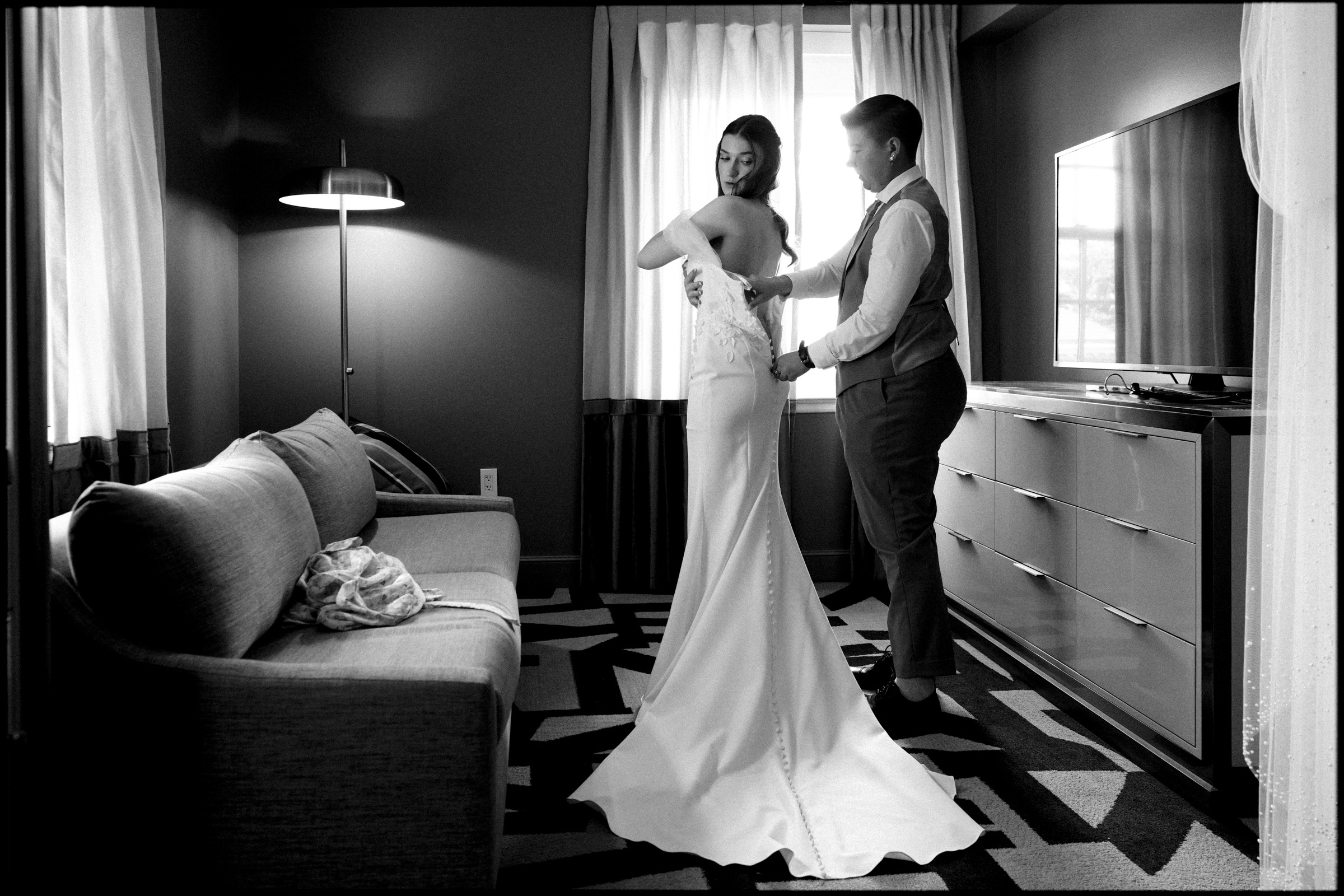 In a modern, softly lit hotel room at the Carolina Inn, a person in a suit helps a bride fasten the back of her gown, capturing an intimate moment before their Elegant Spring Celebration in Chapel Hill. By award-winning Raleigh wedding photographer, Joe Payne.
