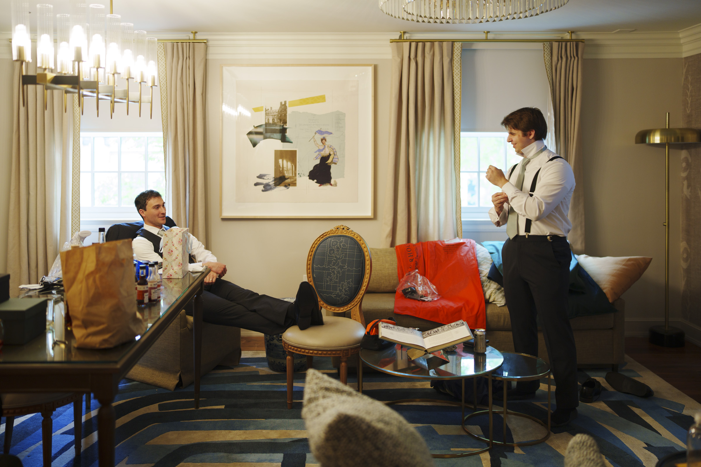 Two men in dress shirts and suspenders talk in a modern living room with snacks, a suit jacket, and ties scattered around. One sits with feet up on a table while the other stands adjusting his cuff, both preparing for an Elegant Spring Celebration at the Carolina Inn Wedding in Chapel Hill. By award-winning Raleigh wedding photographer, Joe Payne.