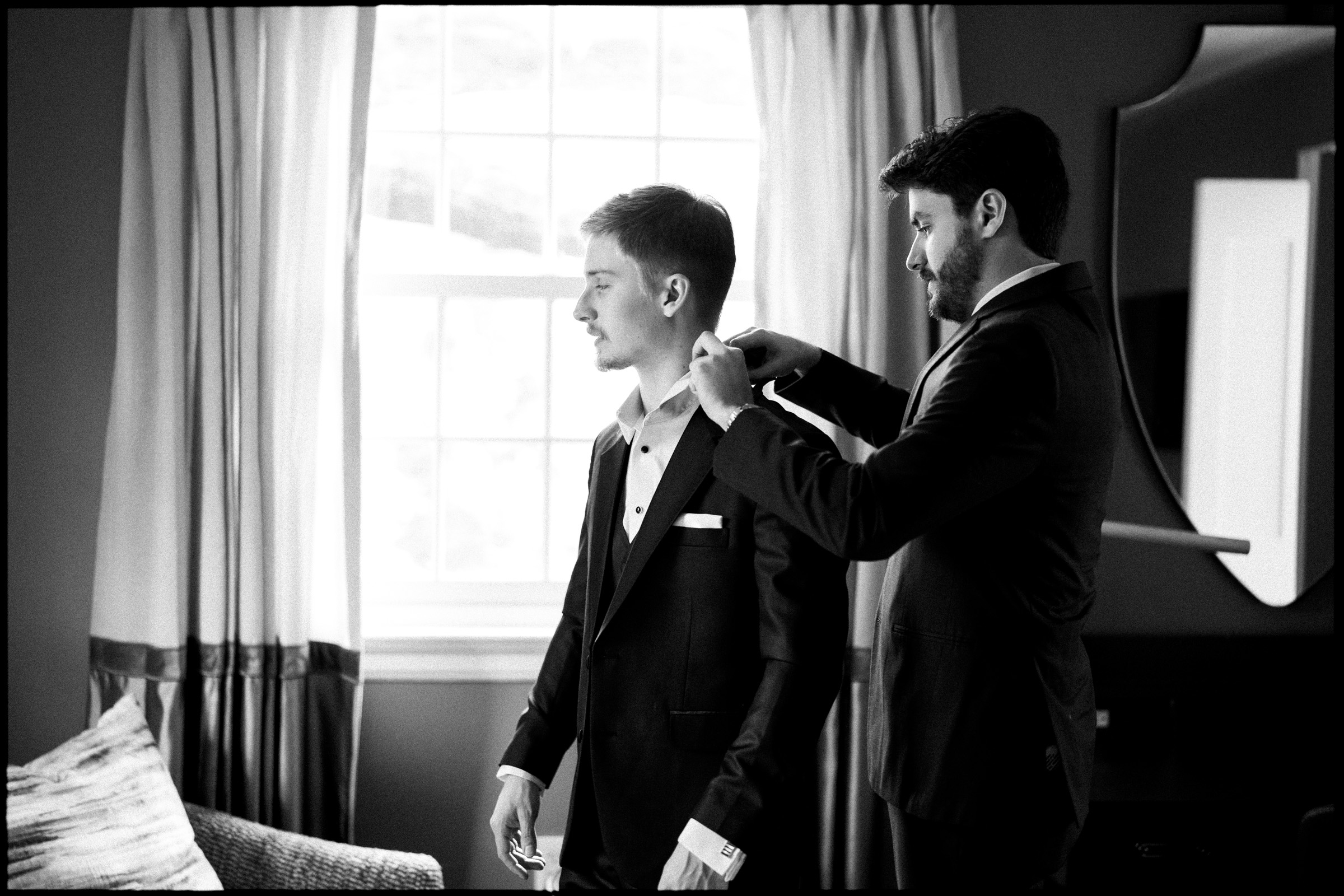 Two men in suits stand in a room with a window at the Carolina Inn, preparing for an Elegant Spring Celebration. One man is helping the other adjust his collar or tie. The scene is black and white, with curtains and a mirror in the background, capturing a quiet moment before the Chapel Hill wedding festivities begin. By award-winning Raleigh wedding photographer, Joe Payne.
