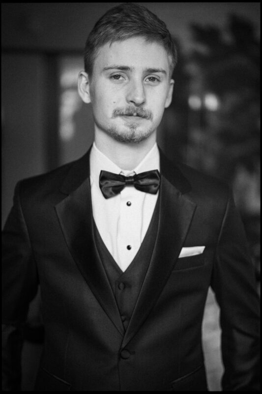 A young man with short hair and a mustache wears a tuxedo with a bow tie, vest, and pocket square. He faces the camera with a neutral expression in this black-and-white photo, capturing the refined atmosphere of an Elegant Spring Celebration at the Carolina Inn Wedding. By award-winning Raleigh wedding photographer, Joe Payne.