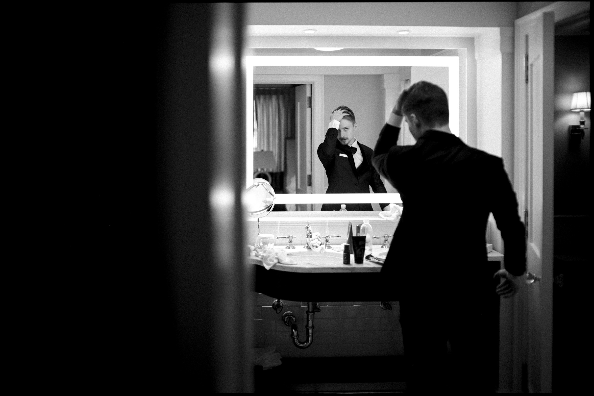 A person in a suit stands at a bathroom sink, adjusting their hair while looking into a large, bright mirror. The scene is shown in black and white, with a clear reflection of the person in the mirror—capturing an elegant moment of preparation before an Elegant Spring Celebration at the Carolina Inn Wedding in Chapel Hill. By award-winning Raleigh wedding photographer, Joe Payne.