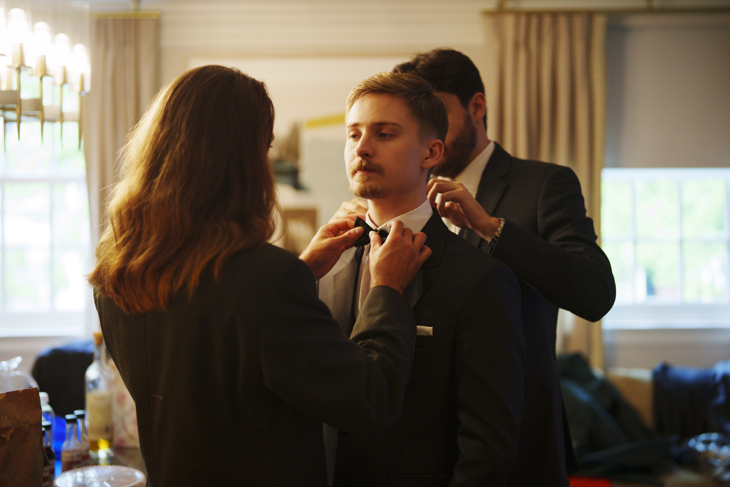 Three men in suits stand indoors as two of them help the third adjust his bow tie. The room is softly lit with curtains and a chandelier, suggesting a formal event or preparation for a special occasion, perhaps an elegant spring celebration at a Carolina Inn Wedding in Chapel Hill. By award-winning Raleigh wedding photographer, Joe Payne.