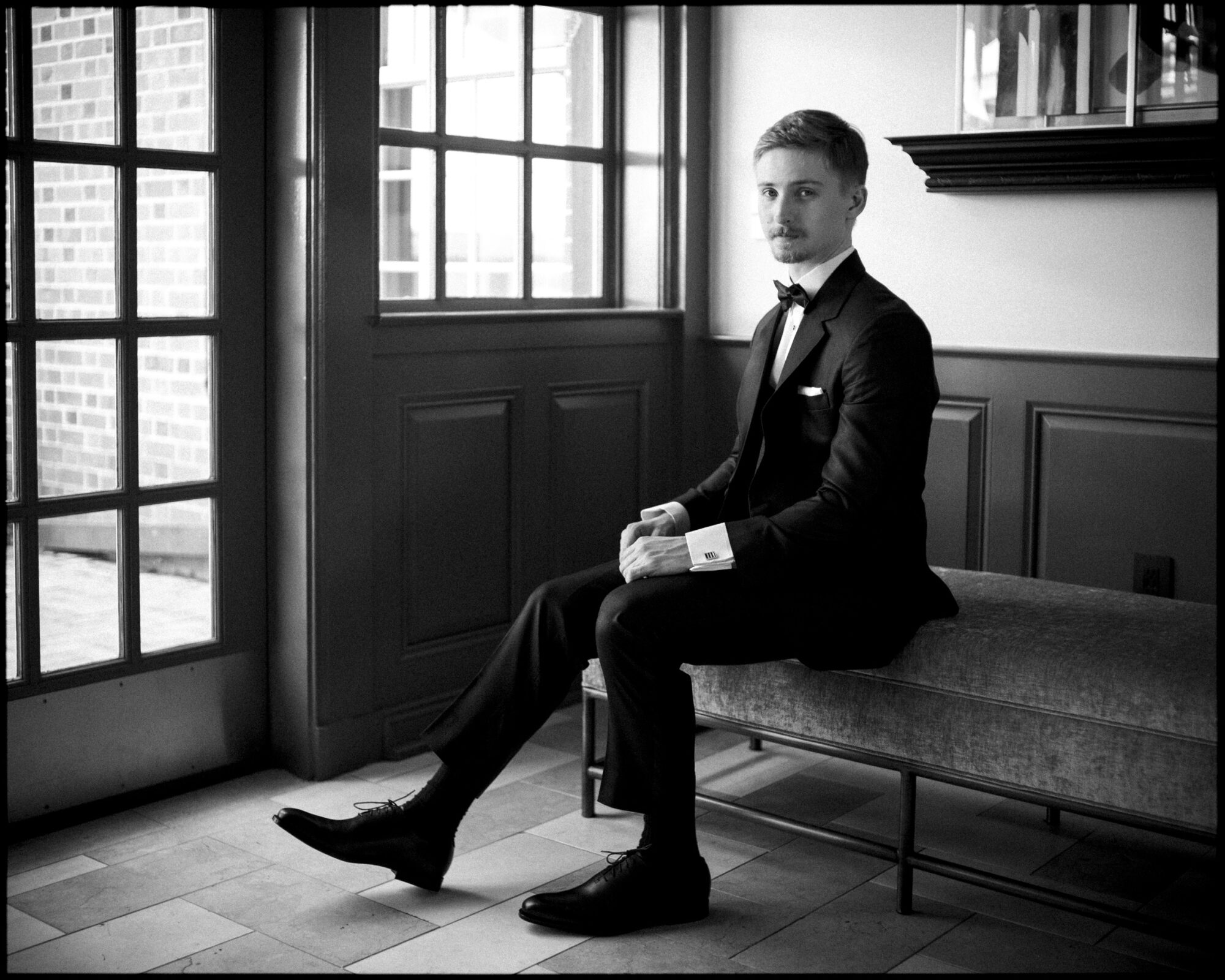 A man in a tuxedo sits on a bench by large windows in a stylish, well-lit room at the Carolina Inn, looking toward the camera with a neutral expression—capturing the timeless atmosphere of this elegant spring celebration in Chapel Hill. By award-winning Raleigh wedding photographer, Joe Payne.