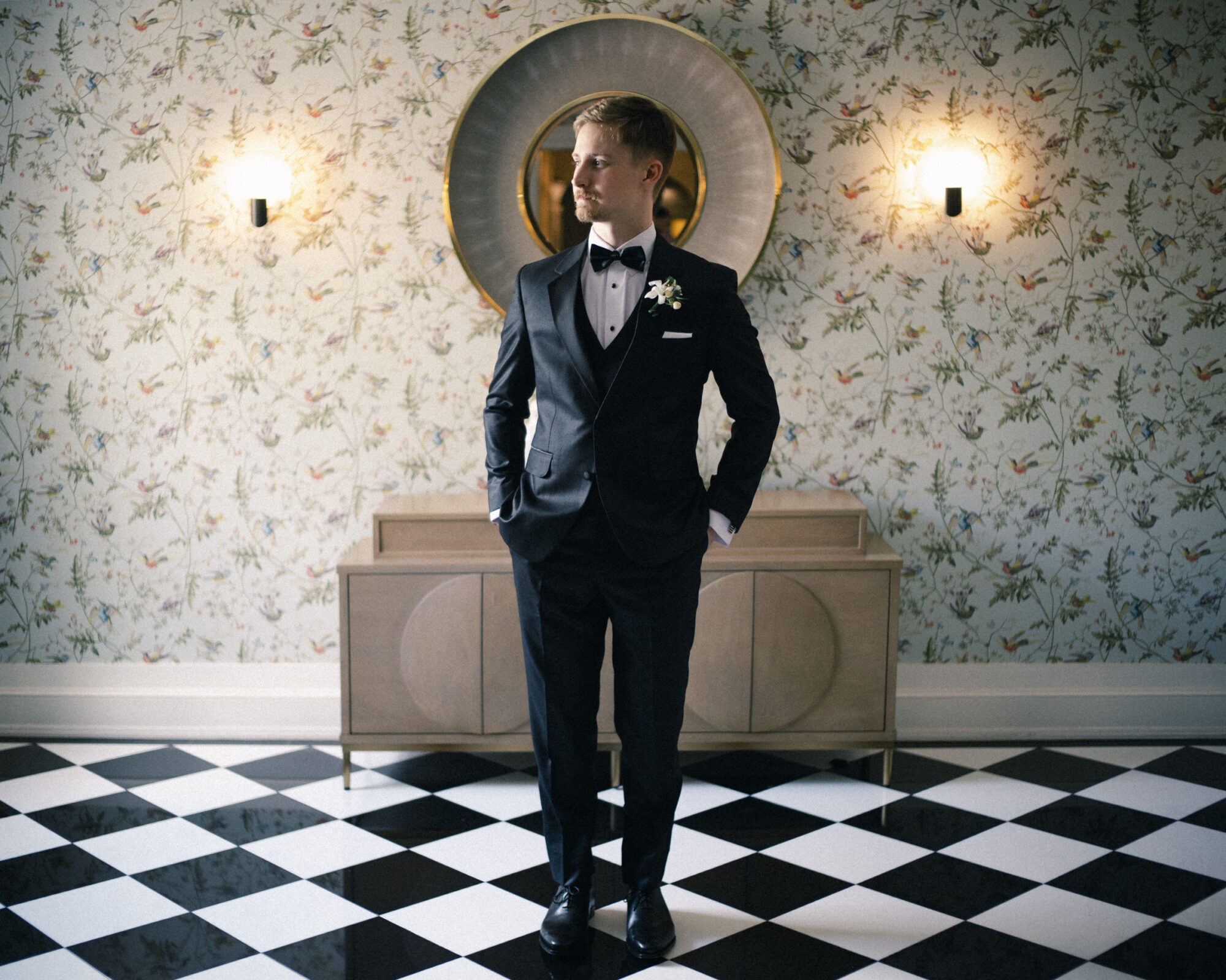 A man in a tuxedo stands on a black-and-white checkered floor, hands in pockets, in front of a floral wallpapered wall with a round mirror and two lit wall sconces—a timeless scene reminiscent of an Elegant Spring Celebration at the Carolina Inn Wedding in Chapel Hill. By award-winning Raleigh wedding photographer, Joe Payne.