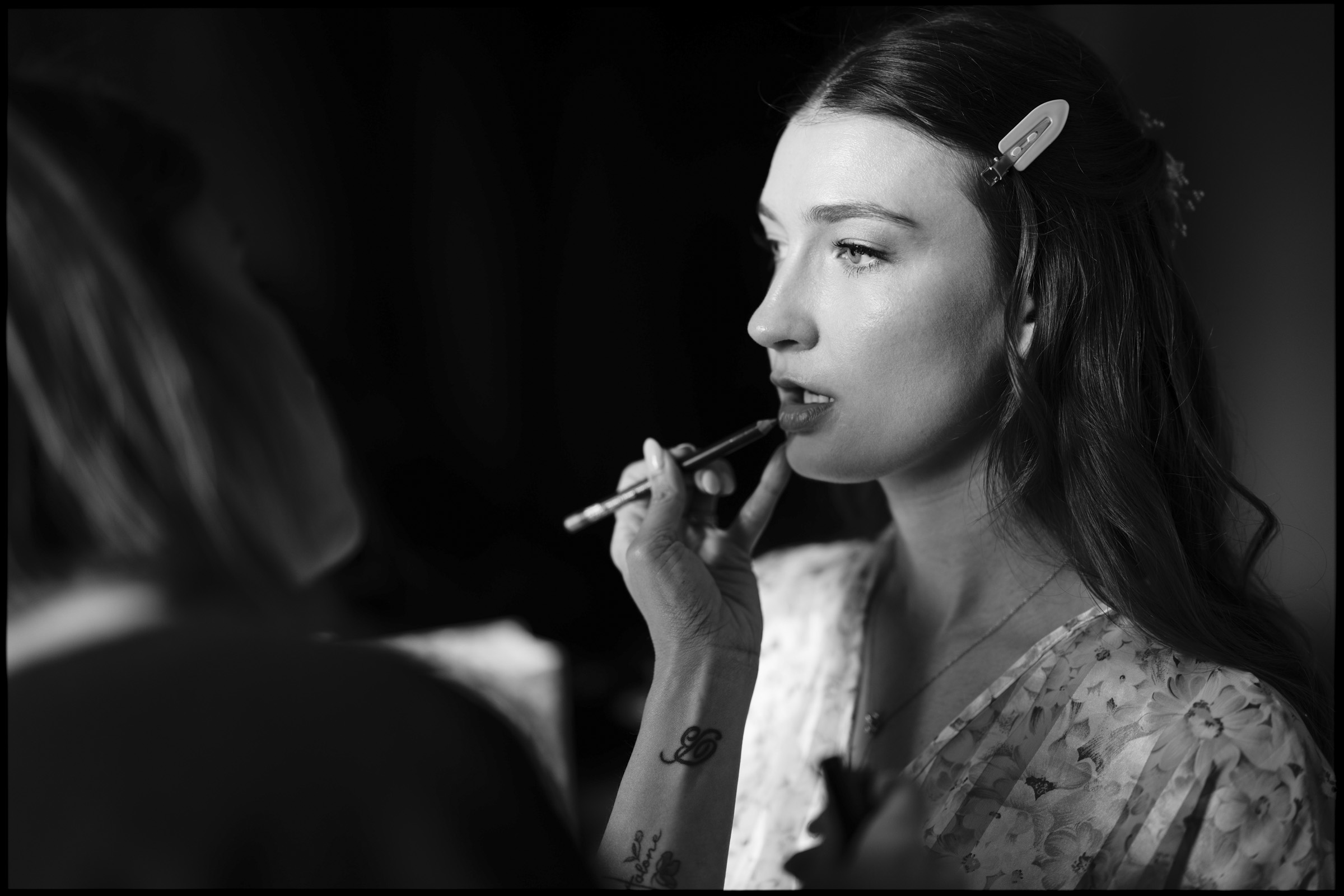 A woman with long hair and a hair clip is having lipstick applied by another person during an Elegant Spring Celebration. The scene is in black and white, and she is wearing a floral top, capturing the timeless charm of a Carolina Inn Wedding in Chapel Hill. By award-winning Raleigh wedding photographer, Joe Payne.