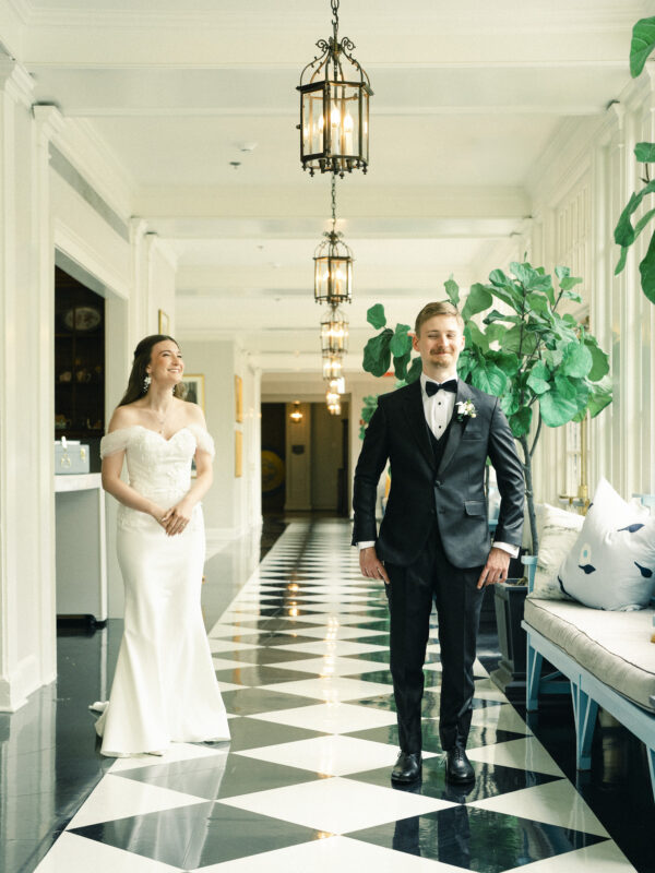 A bride in a white gown smiles at a groom in a black tuxedo as they stand in a bright hallway with black-and-white checkered floors and hanging lanterns, capturing the timeless charm of a Carolina Inn Wedding in Chapel Hill for their elegant spring celebration. By award-winning Raleigh wedding photographer, Joe Payne.