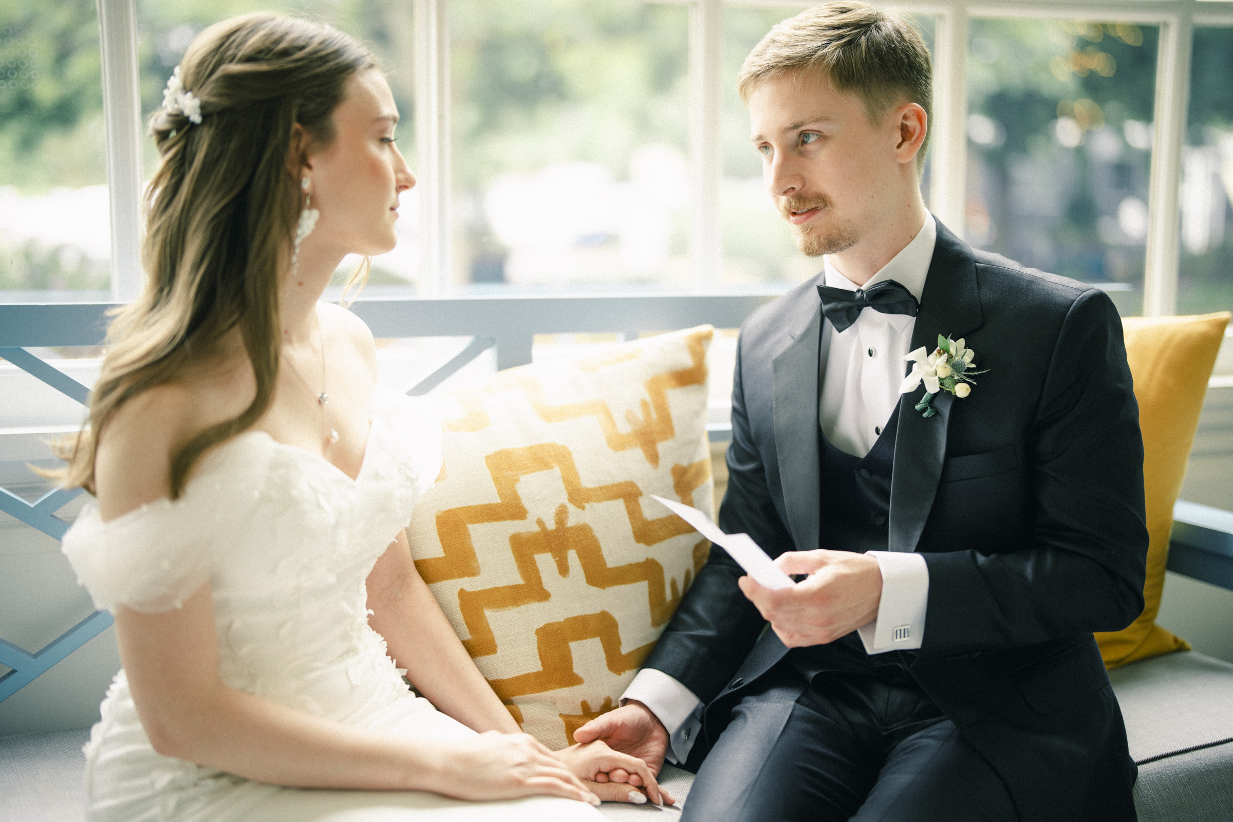 A bride and groom sit together on a bench at their Carolina Inn Wedding in Chapel Hill. The groom, in a black tuxedo, holds a piece of paper and the bride’s hand. The bride wears an off-shoulder white dress. Both look at each other, appearing emotional during this elegant Spring celebration. By award-winning Raleigh wedding photographer, Joe Payne.