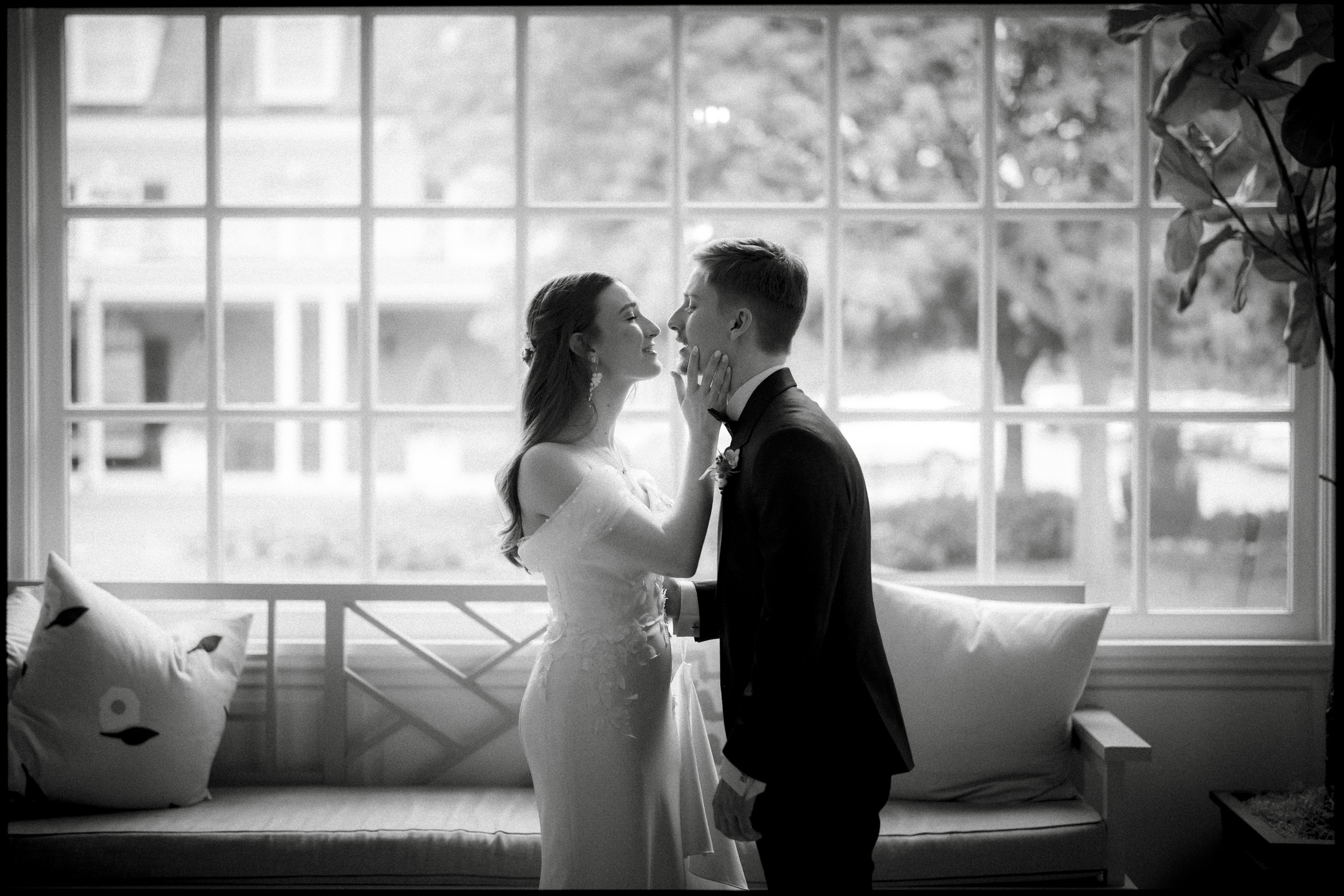 A bride and groom stand close together indoors by a large window at their Carolina Inn Wedding. The bride gently touches the groom’s face as they gaze at each other, both dressed formally. The scene is soft and romantic, with natural light streaming in—a perfect moment from an Elegant Spring Celebration in Chapel Hill. By award-winning Raleigh wedding photographer, Joe Payne.