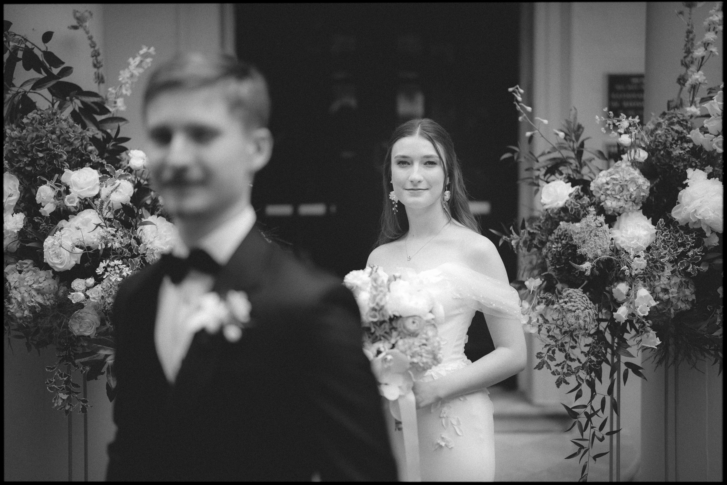 A bride in a white dress holds a bouquet and smiles softly while standing behind a groom in a tuxedo, surrounded by large floral arrangements at an outdoor venue—a scene that perfectly captures the charm of an Elegant Spring Celebration at the Carolina Inn Wedding in Chapel Hill. By award-winning Raleigh wedding photographer, Joe Payne.