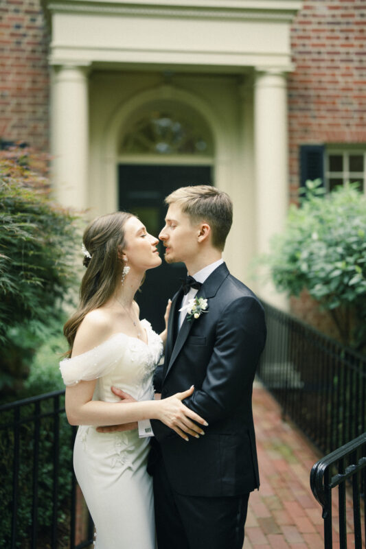 A bride in a white gown and a groom in a black tuxedo stand face-to-face, about to kiss, outside the historic Carolina Inn with its stately brick facade and white columns. The bride’s hand rests on the groom’s waist as they embrace on a brick pathway, capturing the essence of an Elegant Spring Celebration in Chapel Hill. By award-winning Raleigh wedding photographer, Joe Payne.