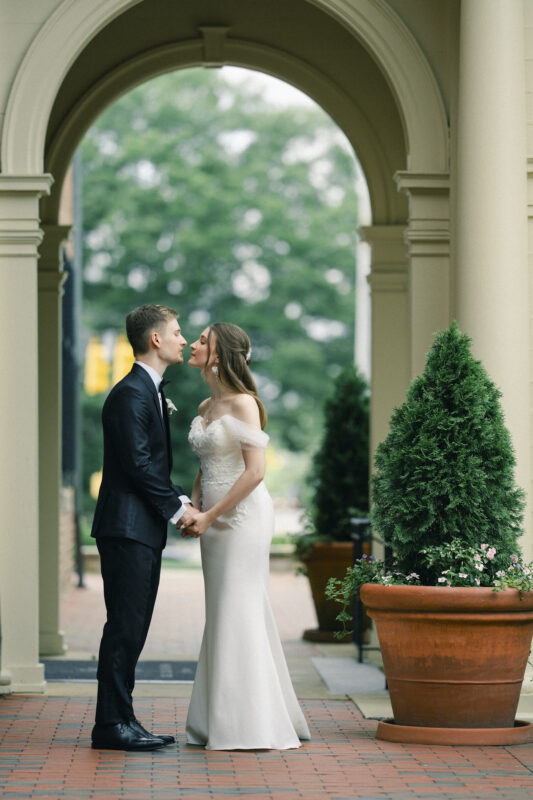A bride and groom stand under an archway at their Elegant Spring Celebration, holding hands and sharing a gentle kiss. The bride wears an off-shoulder white dress, and the groom is in a black suit. Large potted plants frame the scene, capturing the timeless charm of a Carolina Inn Wedding in Chapel Hill. By award-winning Raleigh wedding photographer, Joe Payne.