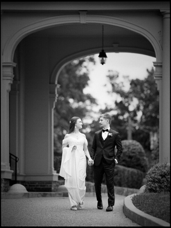 A bride in a long dress and a groom in a tuxedo walk hand in hand under an archway, smiling at each other, with trees and bushes in the background—capturing the timeless romance of an elegant spring celebration at their Carolina Inn wedding. By award-winning Raleigh wedding photographer, Joe Payne.