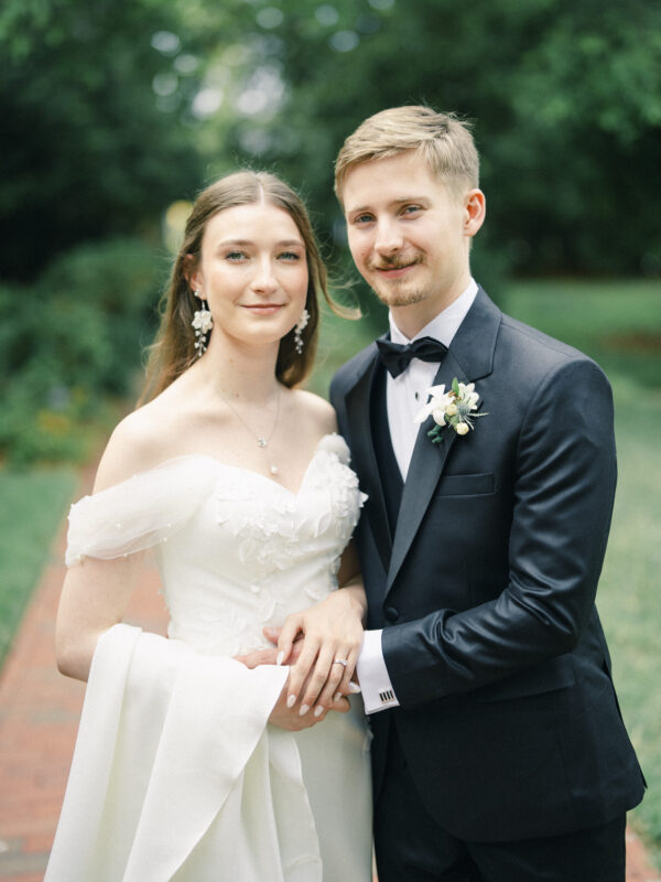 A bride in a white dress and a groom in a black tuxedo stand outdoors, holding hands and smiling at the camera, with lush greenery in the background—a perfect moment from their elegant spring celebration at a Carolina Inn wedding in Chapel Hill. By award-winning Raleigh wedding photographer, Joe Payne.