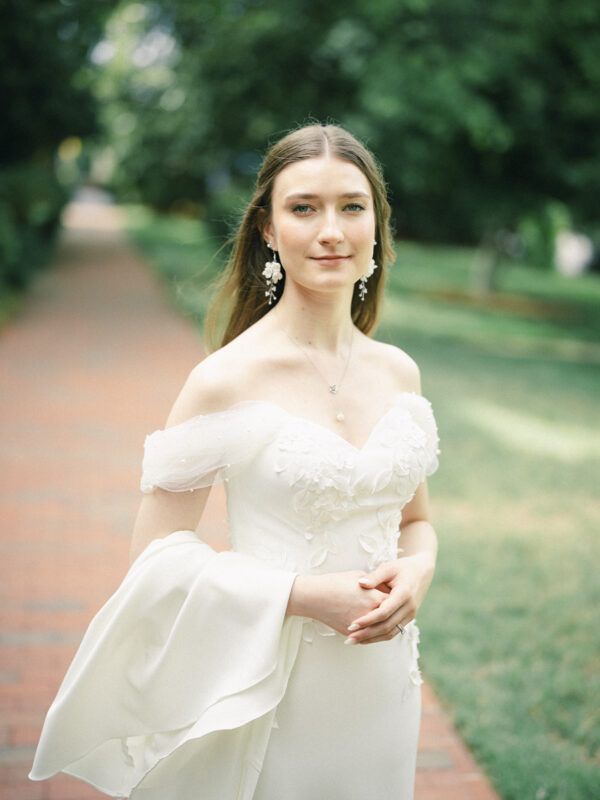 A woman in an off-shoulder white wedding dress stands on a brick path outdoors, surrounded by greenery. She has long brown hair, wears floral earrings, and looks calmly at the camera—perfectly capturing the serene beauty of an Elegant Spring Celebration at the Carolina Inn Wedding in Chapel Hill. By award-winning Raleigh wedding photographer, Joe Payne.