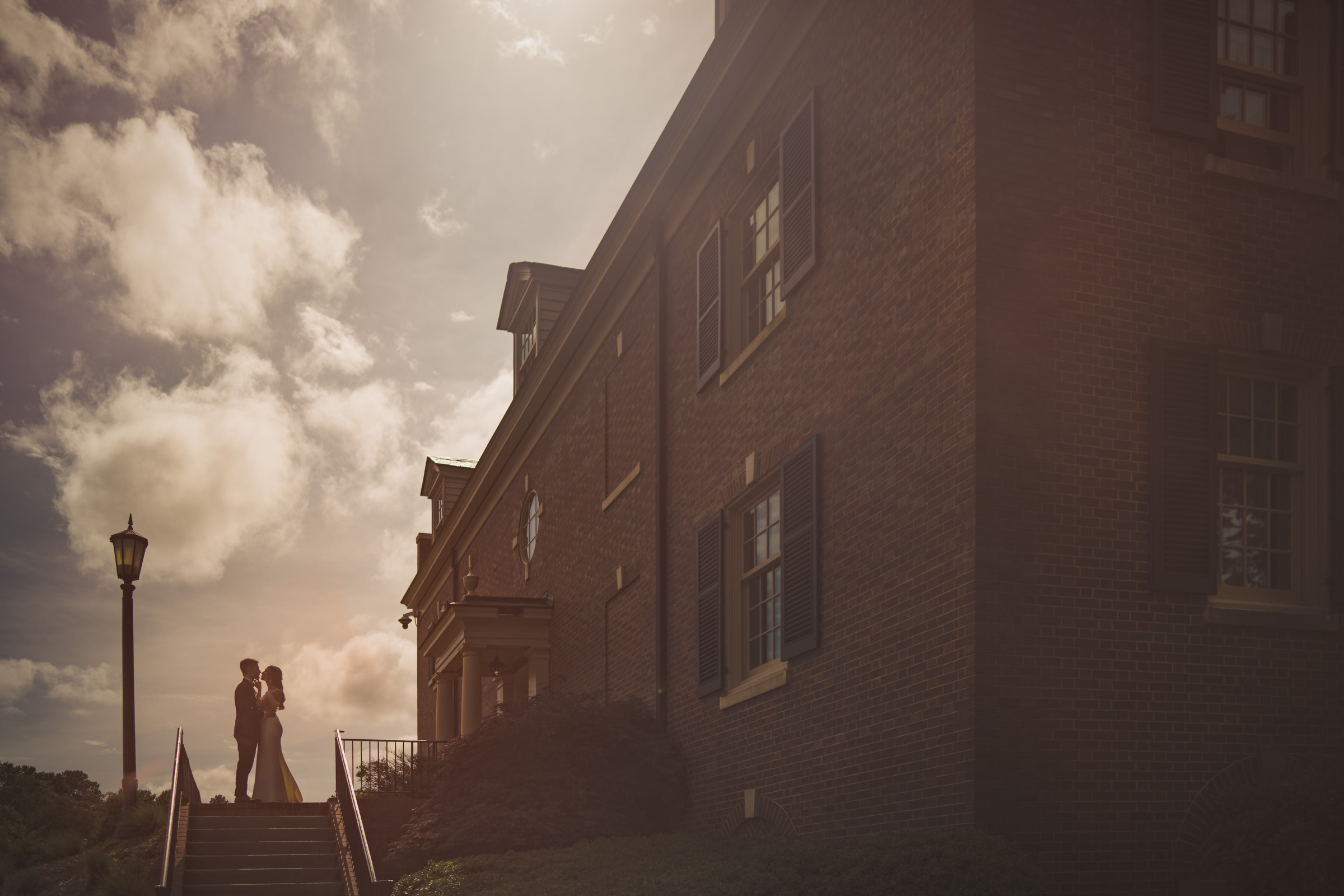 A couple stands at the top of outdoor stairs beside a large brick building, embracing in soft sunlight under a partly cloudy sky—a timeless moment perfectly capturing the romance of an Elegant Spring Celebration at their Carolina Inn Wedding. By award-winning Raleigh wedding photographer, Joe Payne.