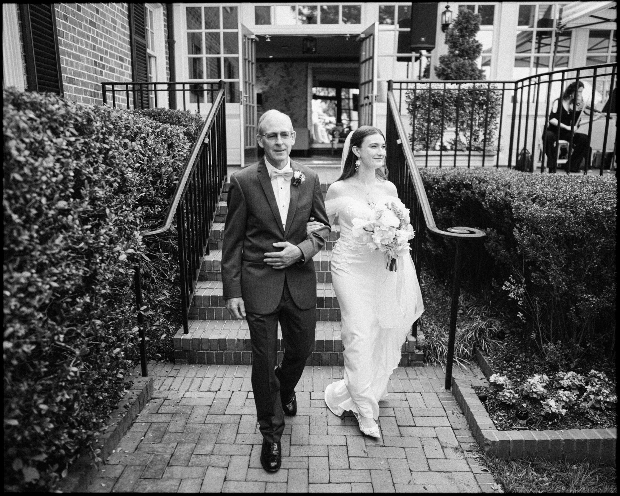 A bride in a white dress holding a bouquet walks arm-in-arm with an older man in a suit down brick steps, outdoors, smiling as they approach the camera during an elegant spring celebration at a Carolina Inn wedding in Chapel Hill. By award-winning Raleigh wedding photographer, Joe Payne.