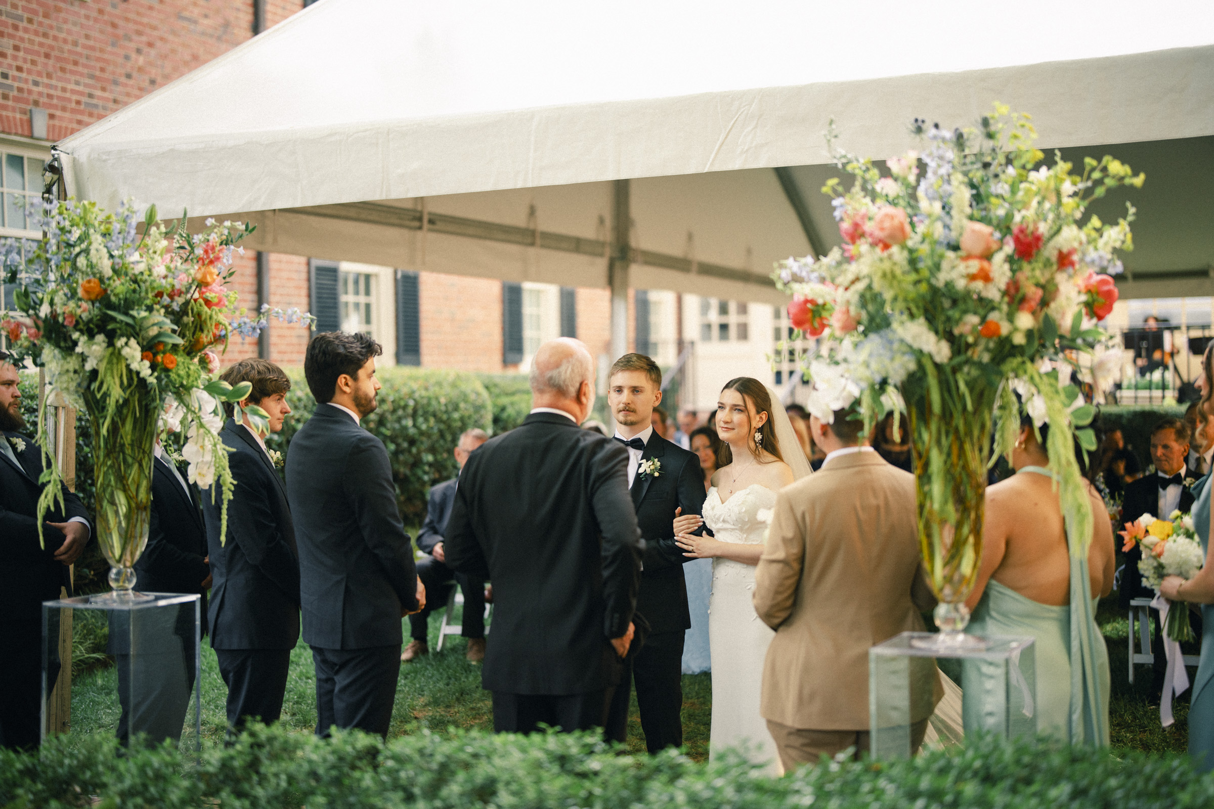 A bride and groom stand together during an elegant spring celebration at a Carolina Inn wedding beneath a white tent, surrounded by guests and large floral arrangements in Chapel Hill. By award-winning Raleigh wedding photographer, Joe Payne.