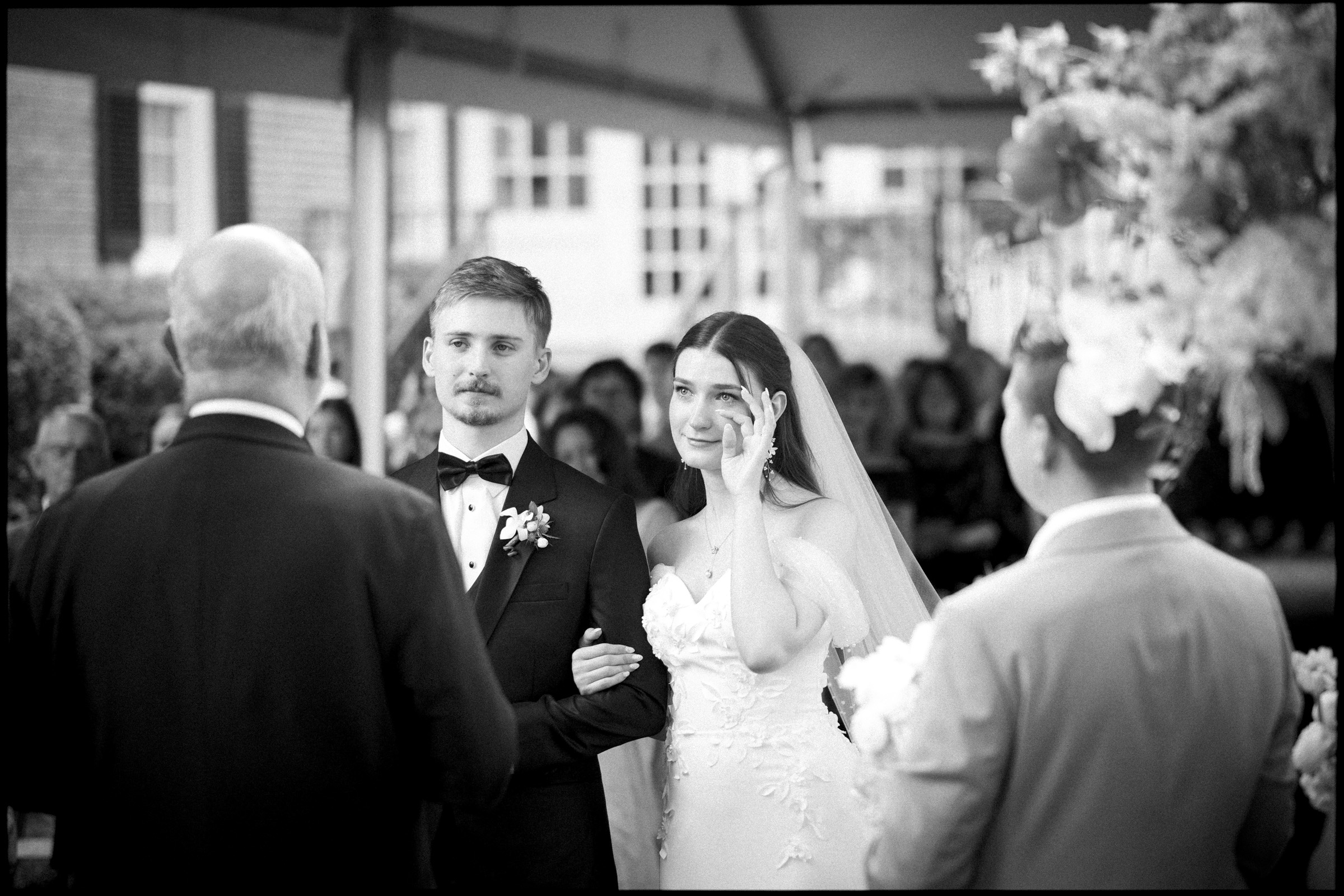 A bride wipes a tear while standing beside the groom during an elegant spring celebration at the Carolina Inn in Chapel Hill, both facing an officiant. The groom looks ahead and holds the bride’s arm as guests sit in the background under a tent. By award-winning Raleigh wedding photographer, Joe Payne.
