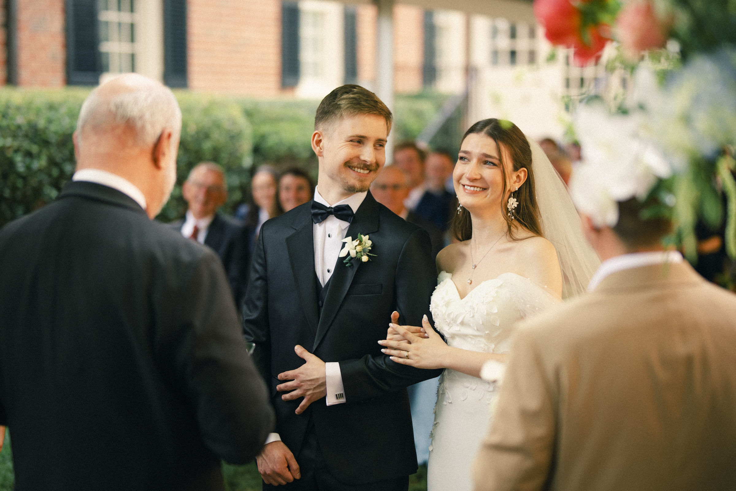 A bride and groom smile while holding hands during an elegant spring celebration at their outdoor Carolina Inn wedding in Chapel Hill, surrounded by guests and floral decorations. By award-winning Raleigh wedding photographer, Joe Payne.