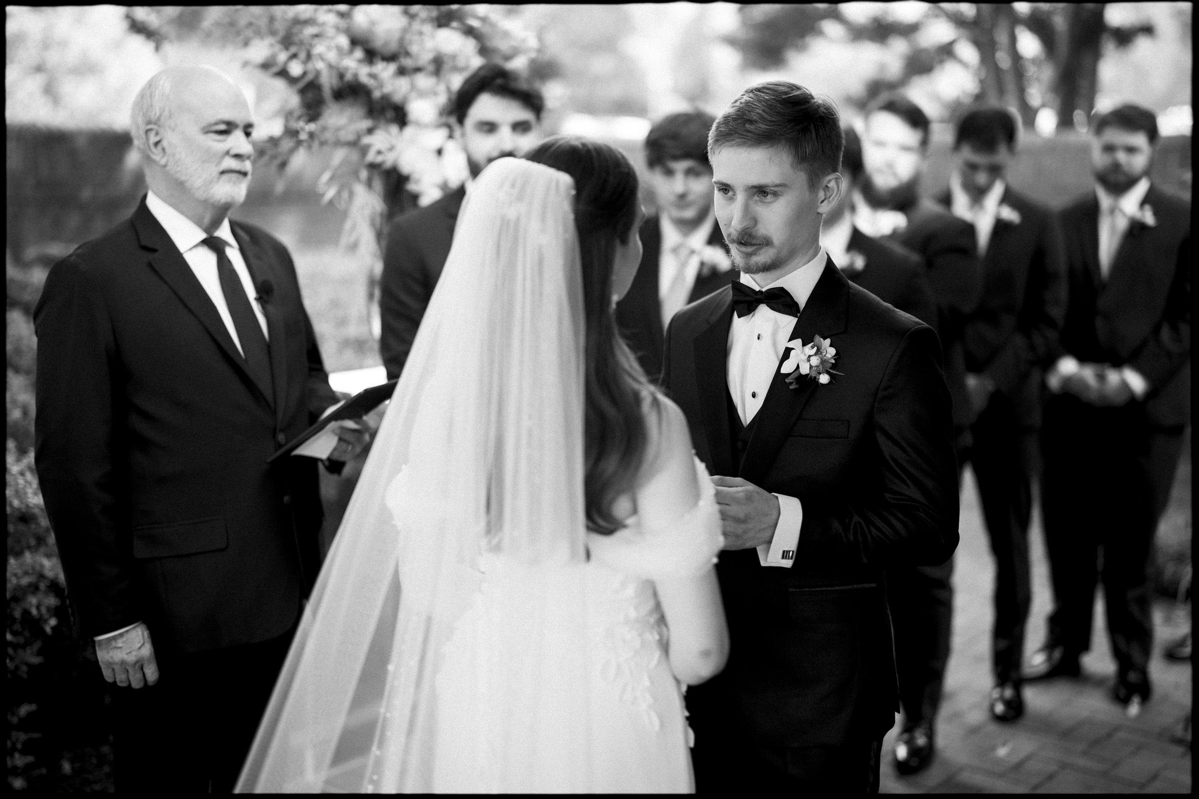 A groom and bride face each other during an outdoor Carolina Inn Wedding ceremony in Chapel Hill, holding hands. An officiant stands nearby, and groomsmen in suits are visible in the background, capturing the essence of an elegant spring celebration. The scene is beautifully rendered in black and white. By award-winning Raleigh wedding photographer, Joe Payne.