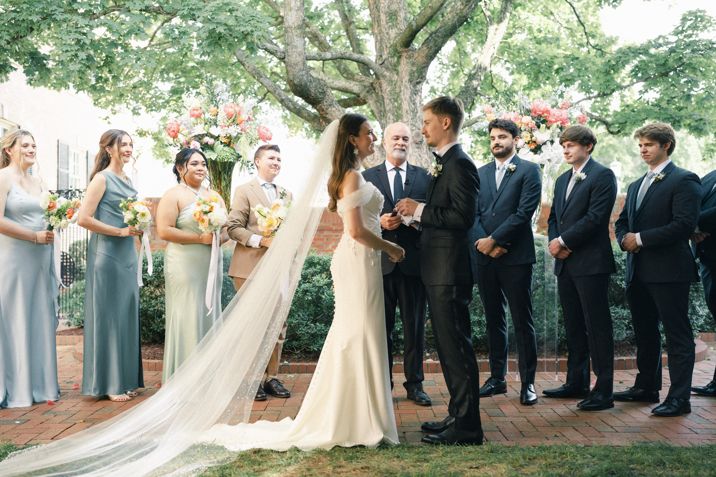 A bride and groom stand facing each other at an outdoor Carolina Inn Wedding ceremony in Chapel Hill, surrounded by their wedding party. Bridesmaids hold bouquets, and a long veil trails behind the bride under a leafy tree, capturing the essence of an elegant spring celebration. By award-winning Raleigh wedding photographer, Joe Payne.
