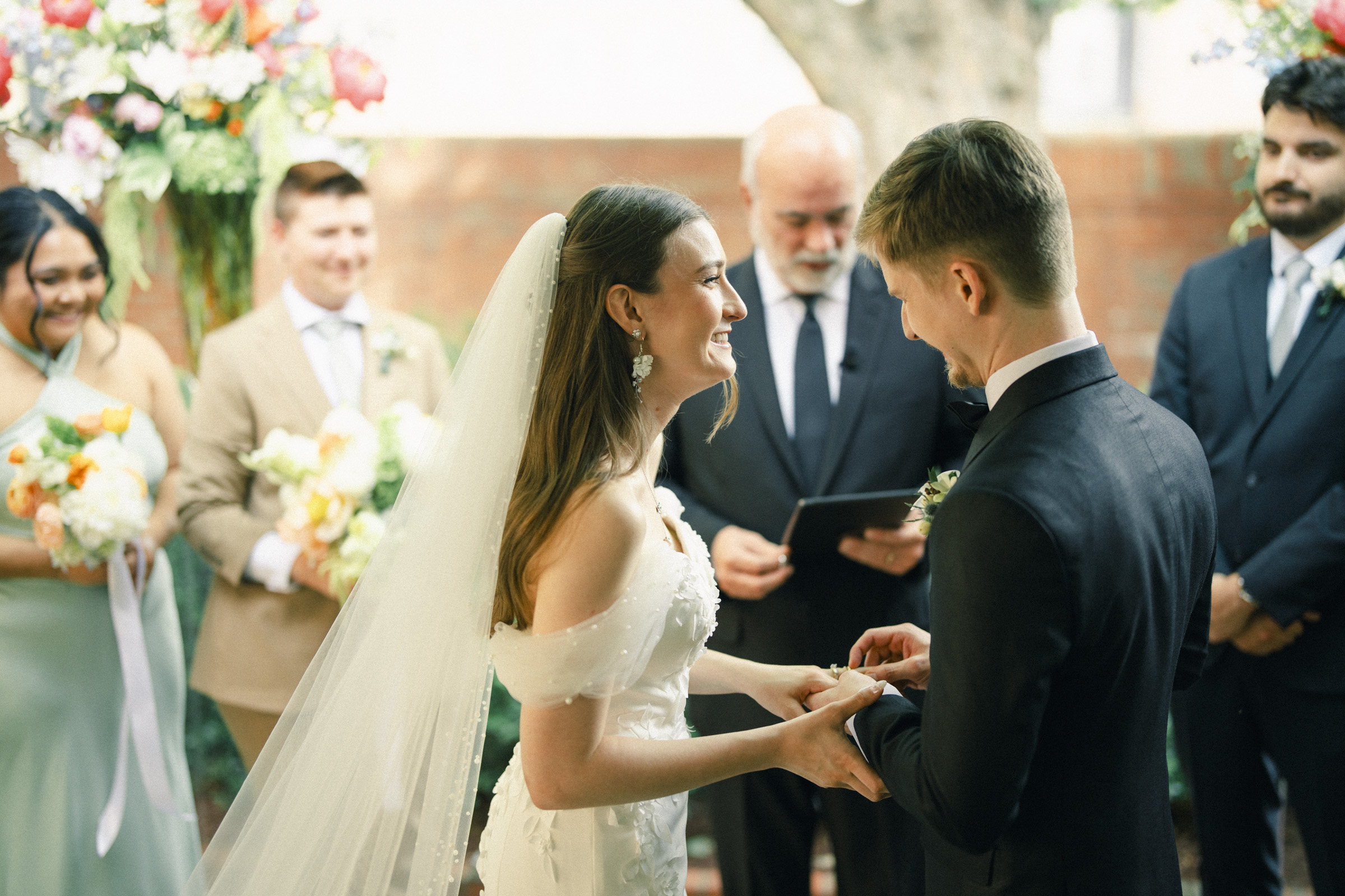 A bride and groom smile at each other while holding hands during their wedding ceremony, surrounded by attendants and an officiant with flowers in the background, capturing the joy of an elegant spring celebration at a beautiful Carolina Inn wedding. By award-winning Raleigh wedding photographer, Joe Payne.