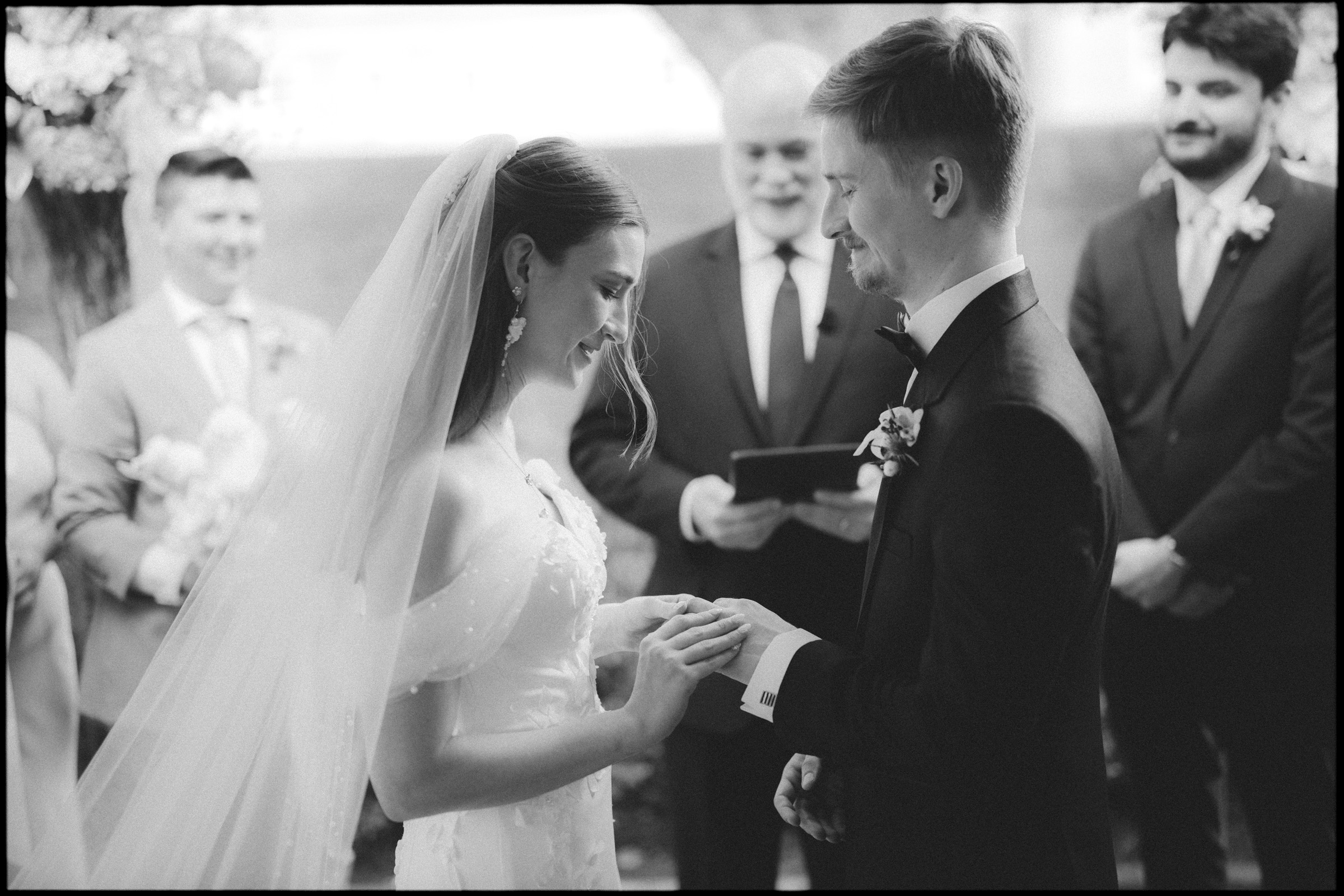 At their elegant spring celebration at the Carolina Inn in Chapel Hill, a bride and groom exchange rings during their wedding ceremony, both smiling. The bride wears a veil and off-shoulder dress; the groom is in a suit. Guests and an officiant stand behind them, witnessing this joyful moment. By award-winning Raleigh wedding photographer, Joe Payne.