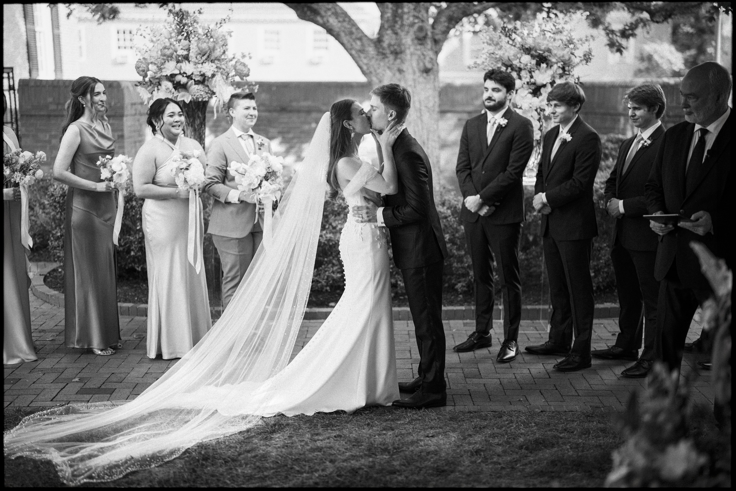 A bride and groom kiss at their elegant spring celebration—a beautiful Carolina Inn wedding in Chapel Hill—surrounded by bridesmaids and groomsmen. The bride wears a long veil, and everyone is smiling and dressed formally. By award-winning Raleigh wedding photographer, Joe Payne.