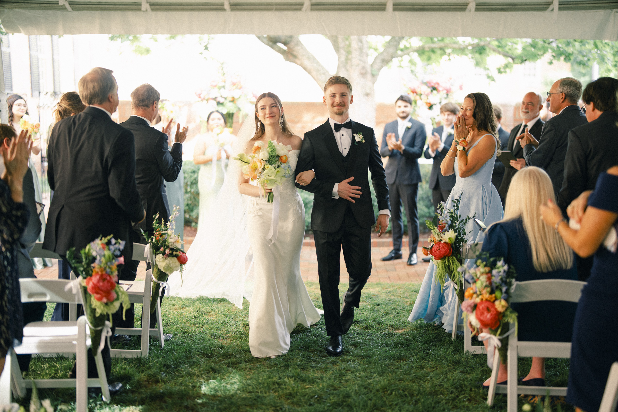 A bride and groom walk arm-in-arm down the aisle outdoors, smiling as guests clap during their Elegant Spring Celebration. The bride holds a bouquet of flowers, and both are dressed in formal wedding attire, creating a timeless moment at their Carolina Inn Wedding. By award-winning Raleigh wedding photographer, Joe Payne.