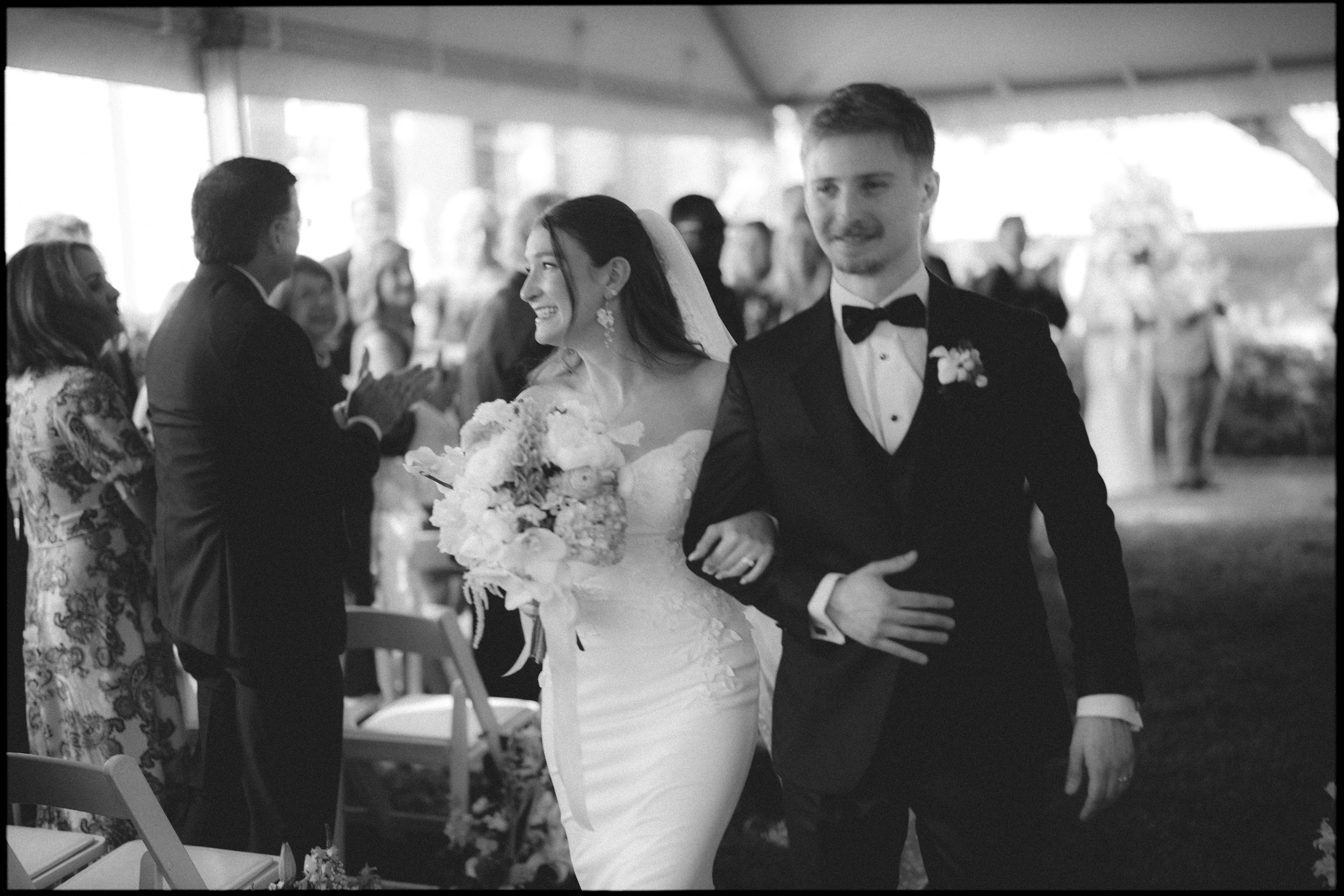 A bride and groom walk arm in arm down the aisle at their Carolina Inn Wedding in Chapel Hill, smiling. The bride holds a bouquet of flowers, while guests seated and standing in the background applaud and watch them during this elegant spring celebration. By award-winning Raleigh wedding photographer, Joe Payne.