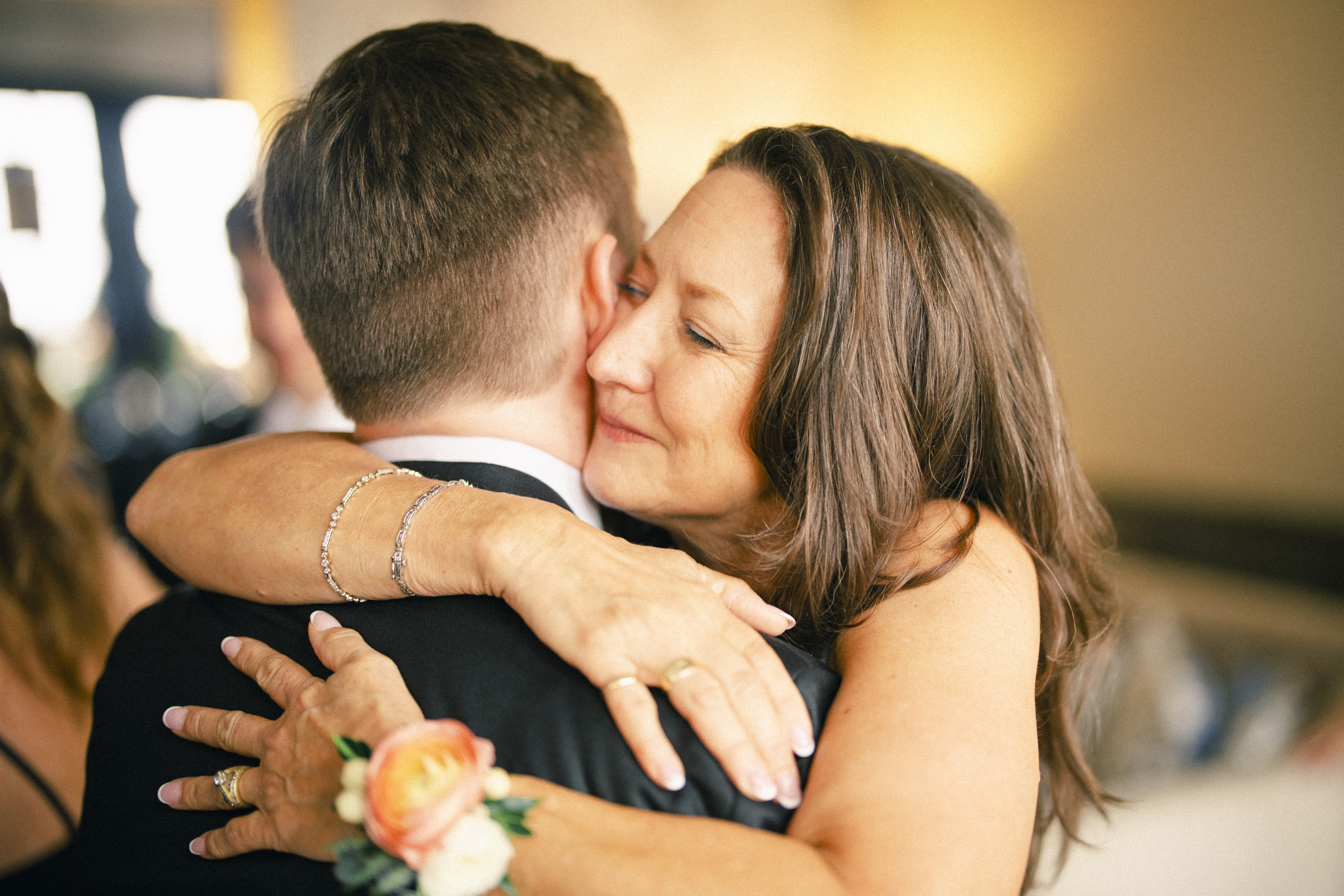 A woman warmly hugs a man in a suit, her eyes closed and smiling. The man wears a boutonniere with peach flowers. The setting is softly lit, suggesting an Elegant Spring Celebration at the Carolina Inn Wedding in Chapel Hill. By award-winning Raleigh wedding photographer, Joe Payne.
