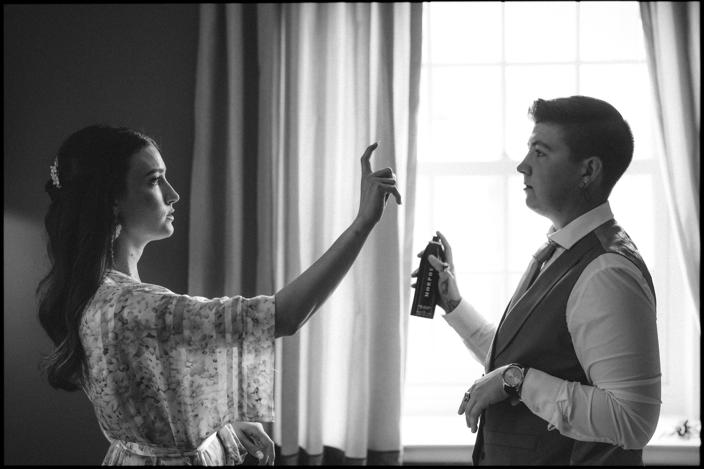 A person in a floral robe sprays hairspray onto another person in a suit, both standing near a window with light streaming in, capturing a quiet moment of preparation before an Elegant Spring Celebration at a Carolina Inn Wedding in Chapel Hill. By award-winning Raleigh wedding photographer, Joe Payne.