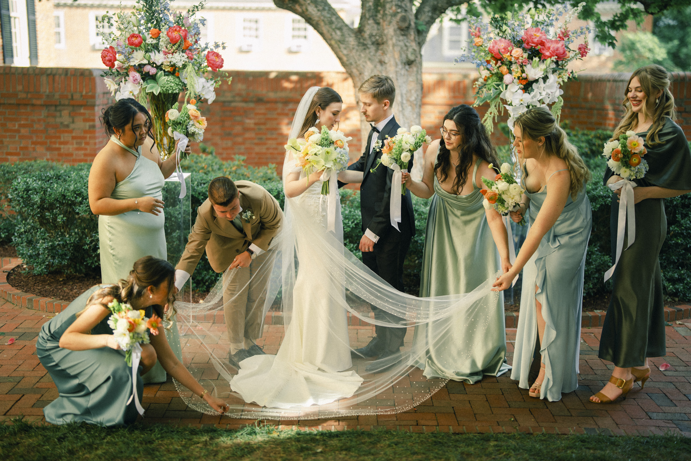 A bride and groom stand outside the Carolina Inn, surrounded by bridesmaids in green dresses who are arranging the bride’s long veil. Colorful flower arrangements frame the group, capturing the essence of an elegant spring celebration in Chapel Hill. By award-winning Raleigh wedding photographer, Joe Payne.