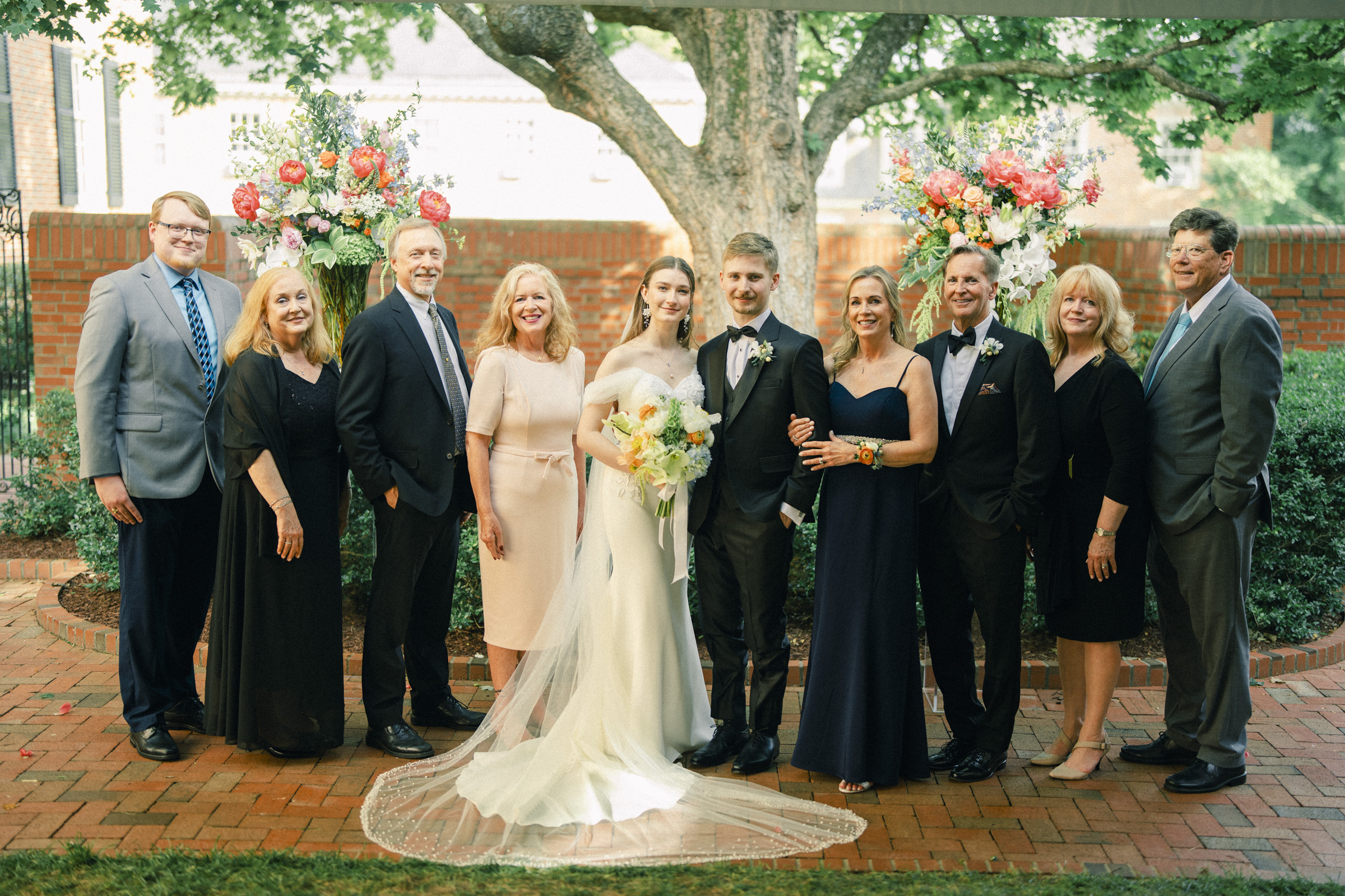 A bride and groom stand outdoors with family and friends in formal attire, surrounded by flowers and greenery on a brick patio at their Carolina Inn Wedding in Chapel Hill, all smiling at the camera during this elegant spring celebration. By award-winning Raleigh wedding photographer, Joe Payne.