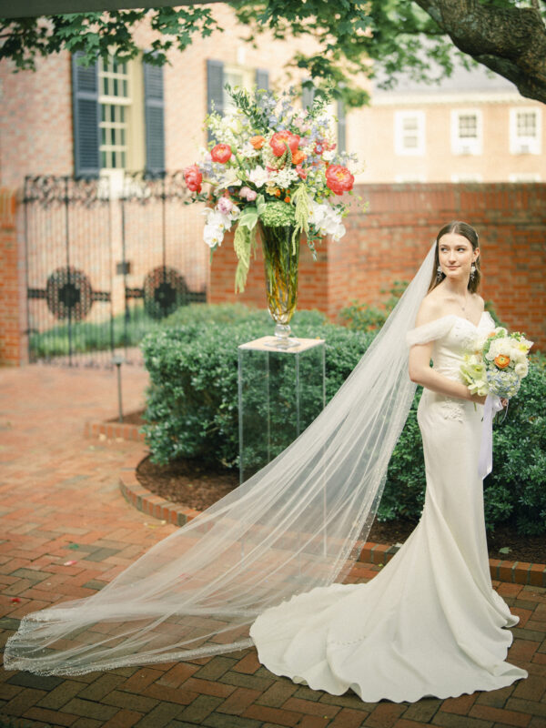 A bride in an elegant white gown with a long veil stands outdoors on a brick patio at the Carolina Inn, holding a bouquet. A large floral arrangement sits on a tall stand beside her, with lush greenery and the charming brick buildings of Chapel Hill in the background—a picture-perfect scene for an elegant spring celebration. By award-winning Raleigh wedding photographer, Joe Payne.