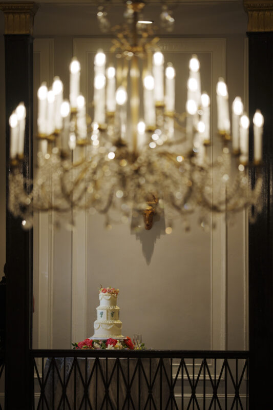 A tiered wedding cake decorated with flowers stands on a table, seen through a softly focused, ornate chandelier hanging from the ceiling—an exquisite detail of this Carolina Inn Wedding, capturing the essence of an elegant spring celebration in Chapel Hill. By award-winning Raleigh wedding photographer, Joe Payne.