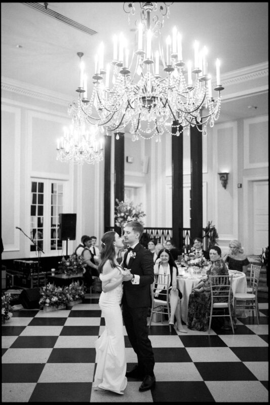A bride and groom share their first dance in an elegant ballroom with chandeliers, checkered floors, and seated guests watching nearby, capturing the timeless romance of a Carolina Inn Wedding. By award-winning Raleigh wedding photographer, Joe Payne.