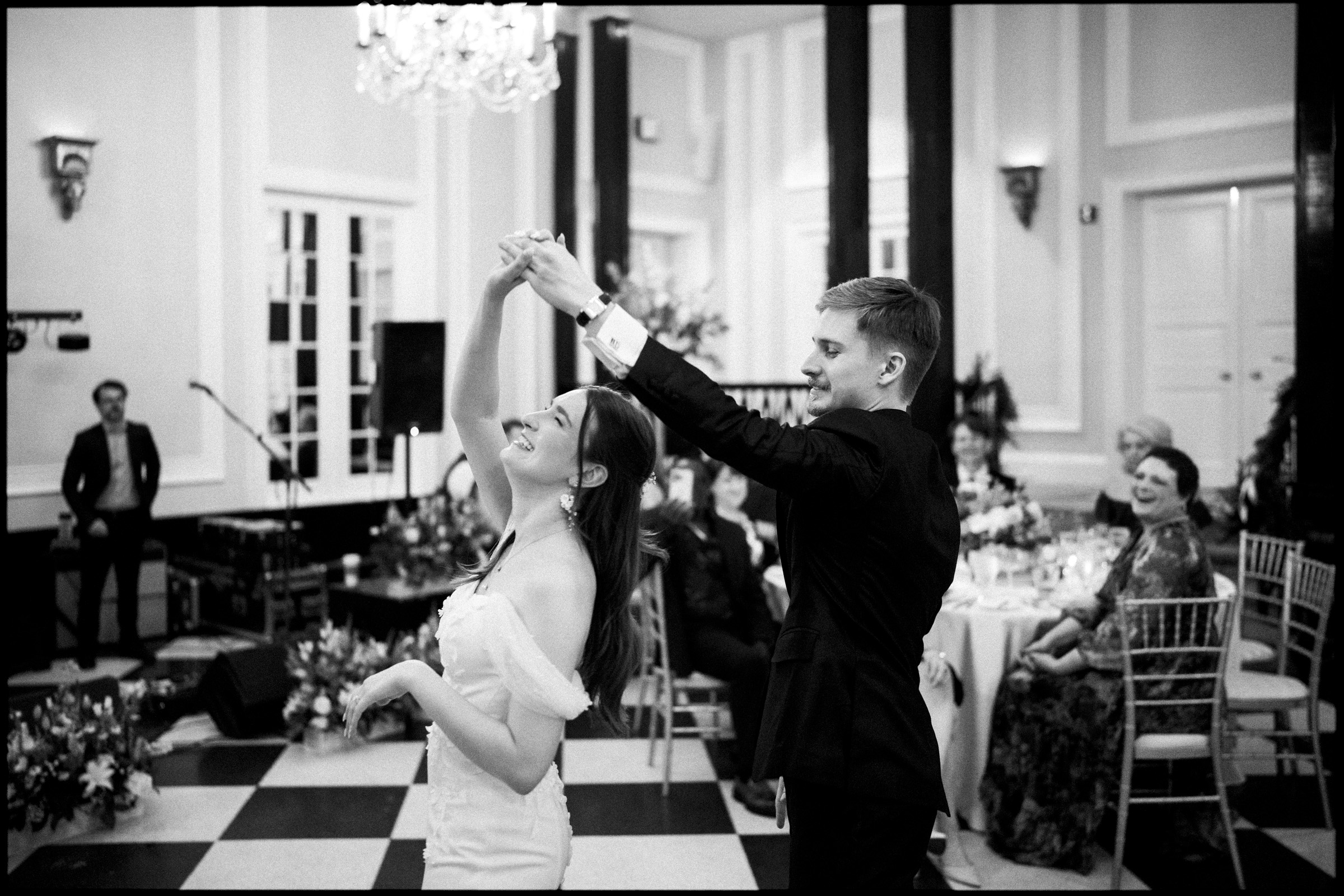 At a Carolina Inn Wedding, a couple dances in a grand ballroom at a formal event, with the woman smiling as the man twirls her. Seated guests watch, surrounded by elegant decor and lush floral arrangements, capturing the charm of an elegant spring wedding. By award-winning Raleigh wedding photographer, Joe Payne.