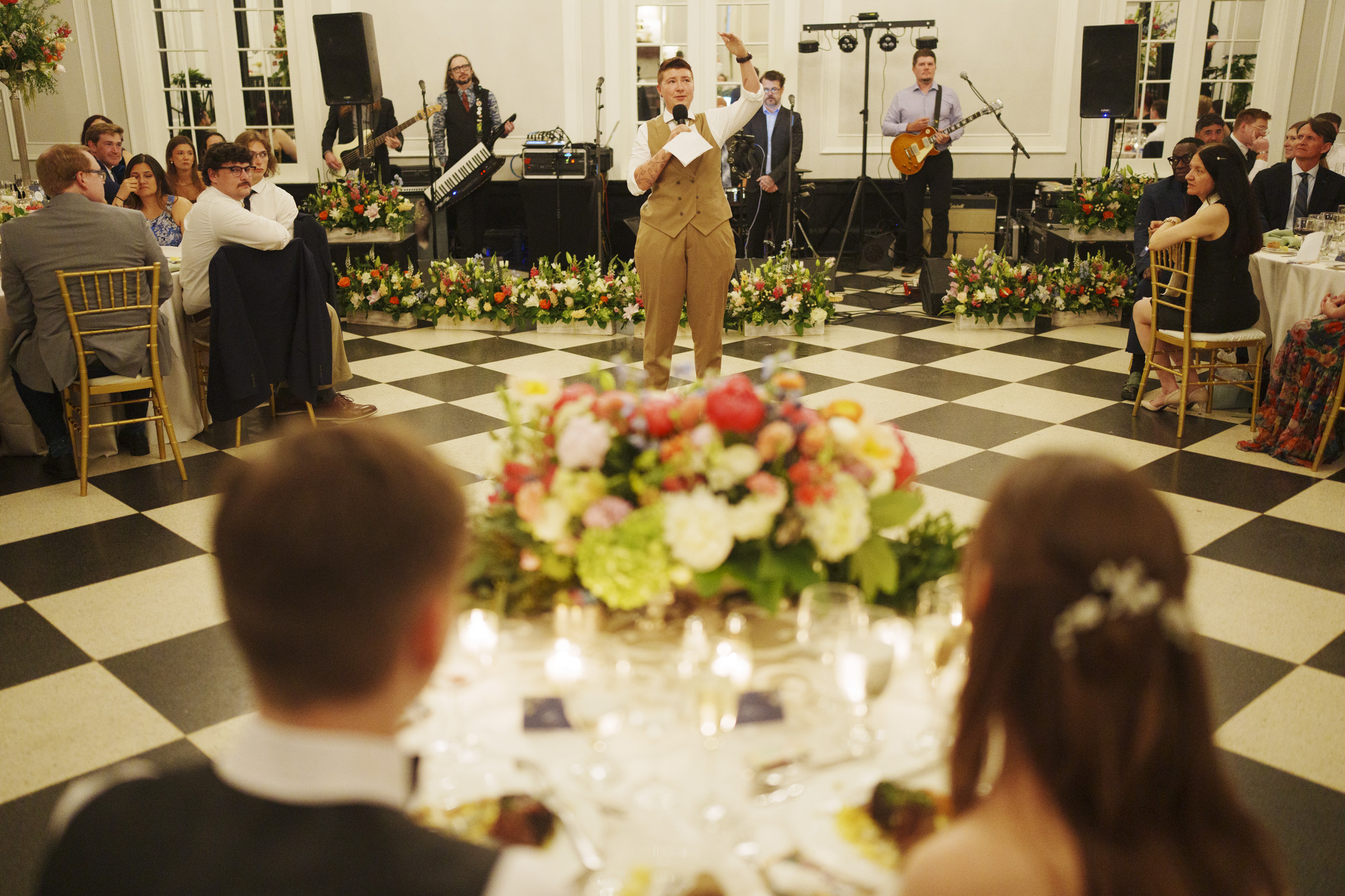A person in a tan suit gives a speech at a wedding reception, with guests seated at round tables, a live band in the background, and large floral centerpieces on checkered floors—capturing the timeless charm of a Carolina Inn Wedding in Chapel Hill during an elegant spring celebration. By award-winning Raleigh wedding photographer, Joe Payne.