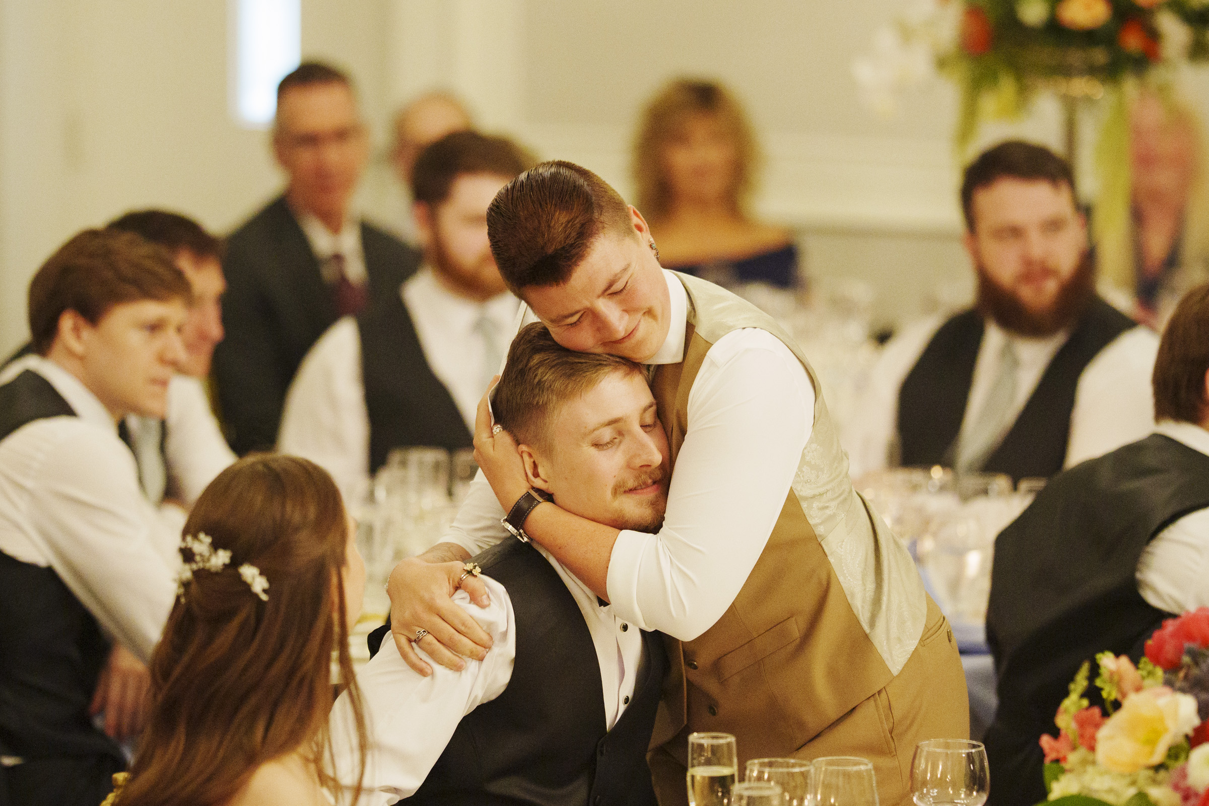Two people embrace warmly at a formal event, surrounded by seated guests dressed in suits and dresses. The scene captures the joy of a Carolina Inn Wedding in Chapel Hill, with tables set with glasses, flowers, and decorations, creating an elegant spring celebration and a truly festive atmosphere. By award-winning Raleigh wedding photographer, Joe Payne.