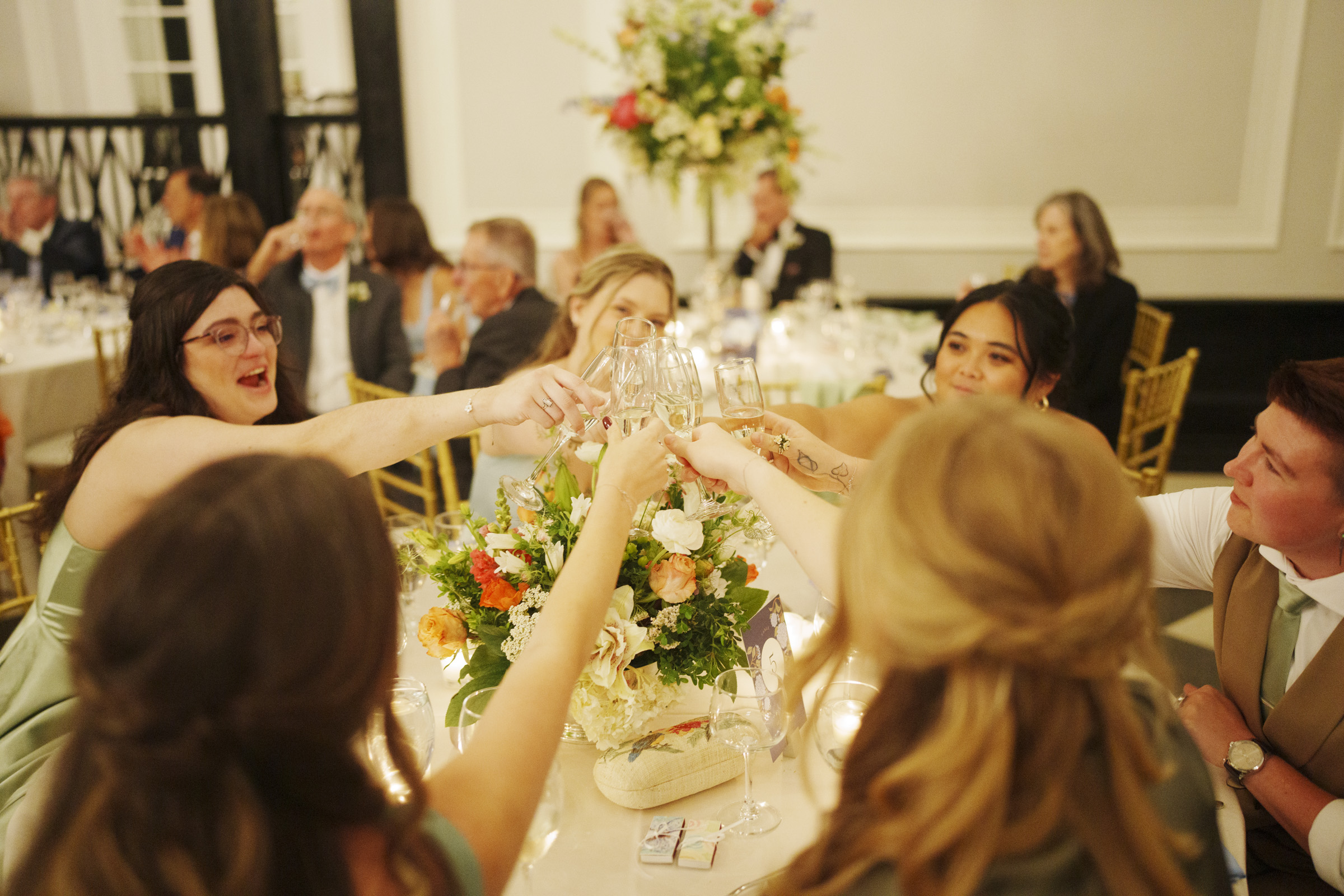 A group of people dressed formally sit around a table at a Carolina Inn Wedding in Chapel Hill, raising their glasses in a toast. The table is decorated with flowers and candles, creating an elegant spring celebration atmosphere, while others are seated in the background. By award-winning Raleigh wedding photographer, Joe Payne.