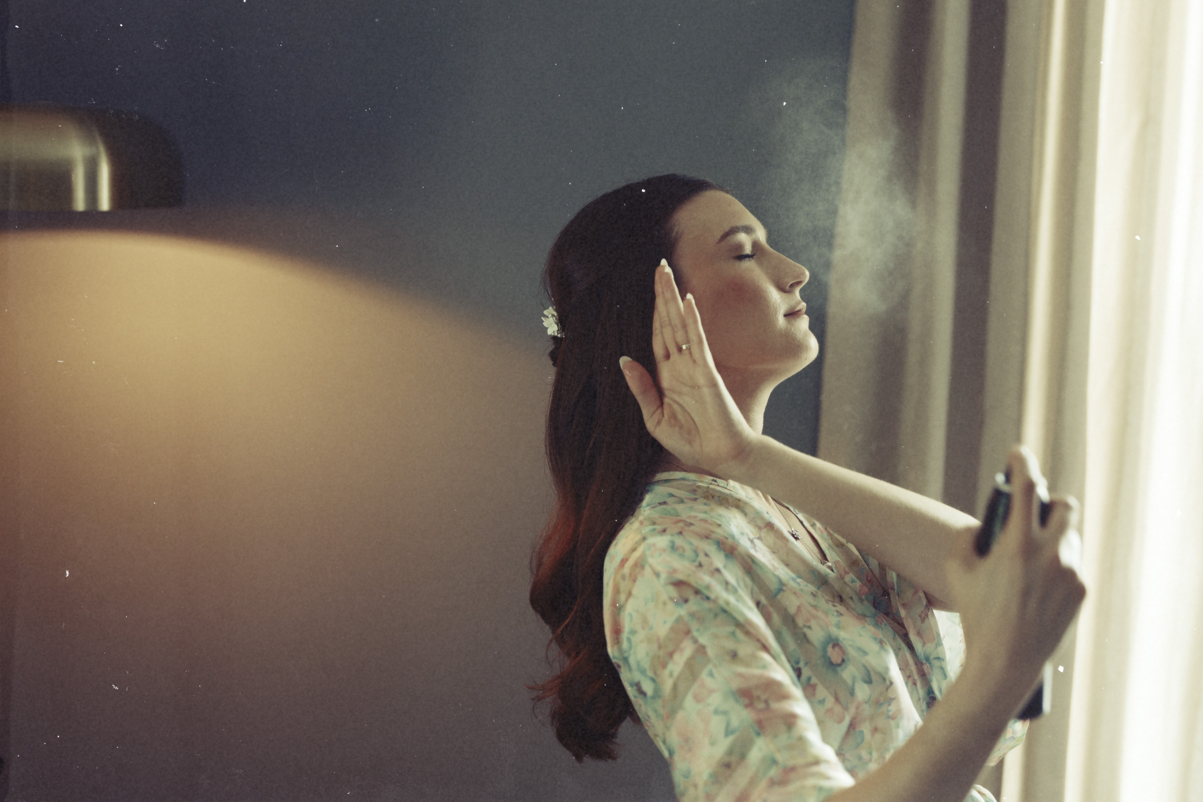 A woman in a floral robe sprays mist on her face, eyes closed and hand raised, standing near a window with soft light coming through the curtains—a serene moment of preparation before an Elegant Spring Celebration at the Carolina Inn Wedding in Chapel Hill. By award-winning Raleigh wedding photographer, Joe Payne.