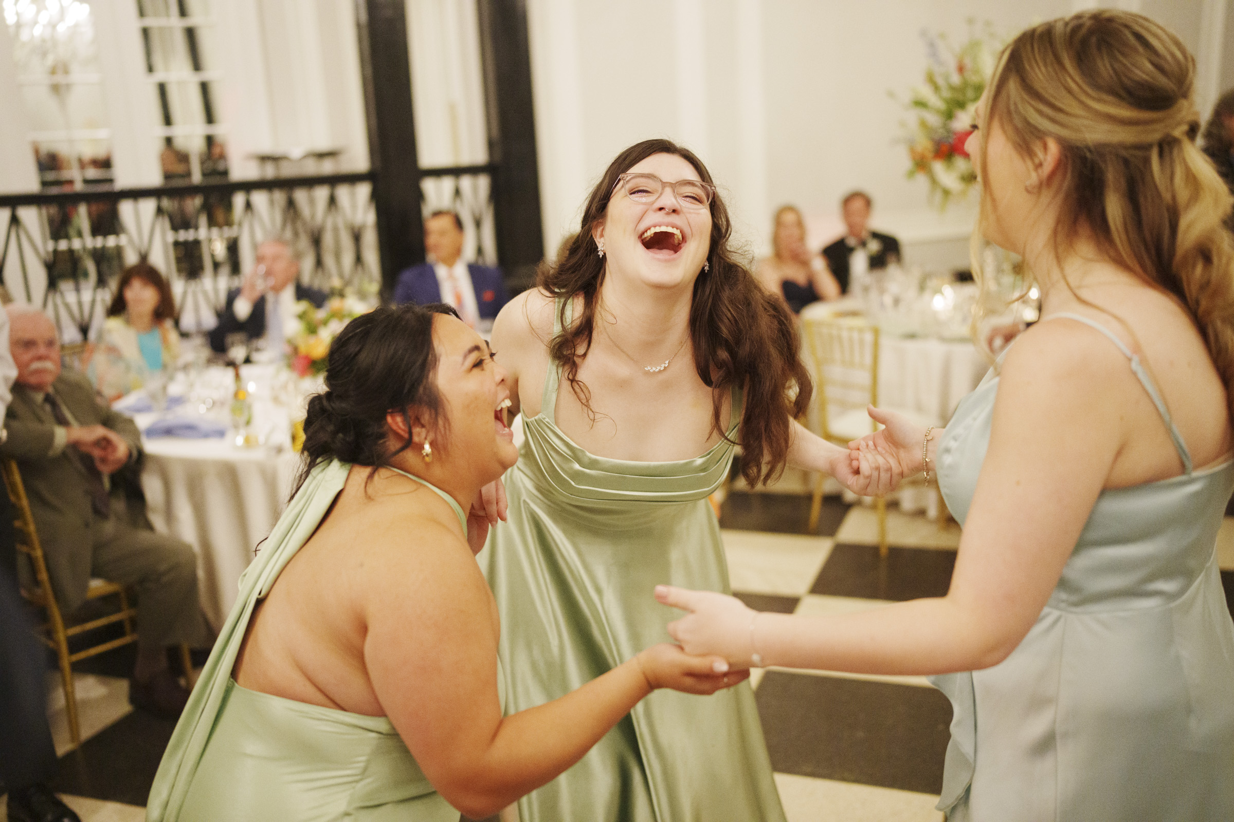 Three women in light green dresses laugh and hold hands as they dance together at a formal event, with seated guests watching and decorated tables in the background—a joyful moment from an elegant spring celebration at a Carolina Inn wedding in Chapel Hill. By award-winning Raleigh wedding photographer, Joe Payne.