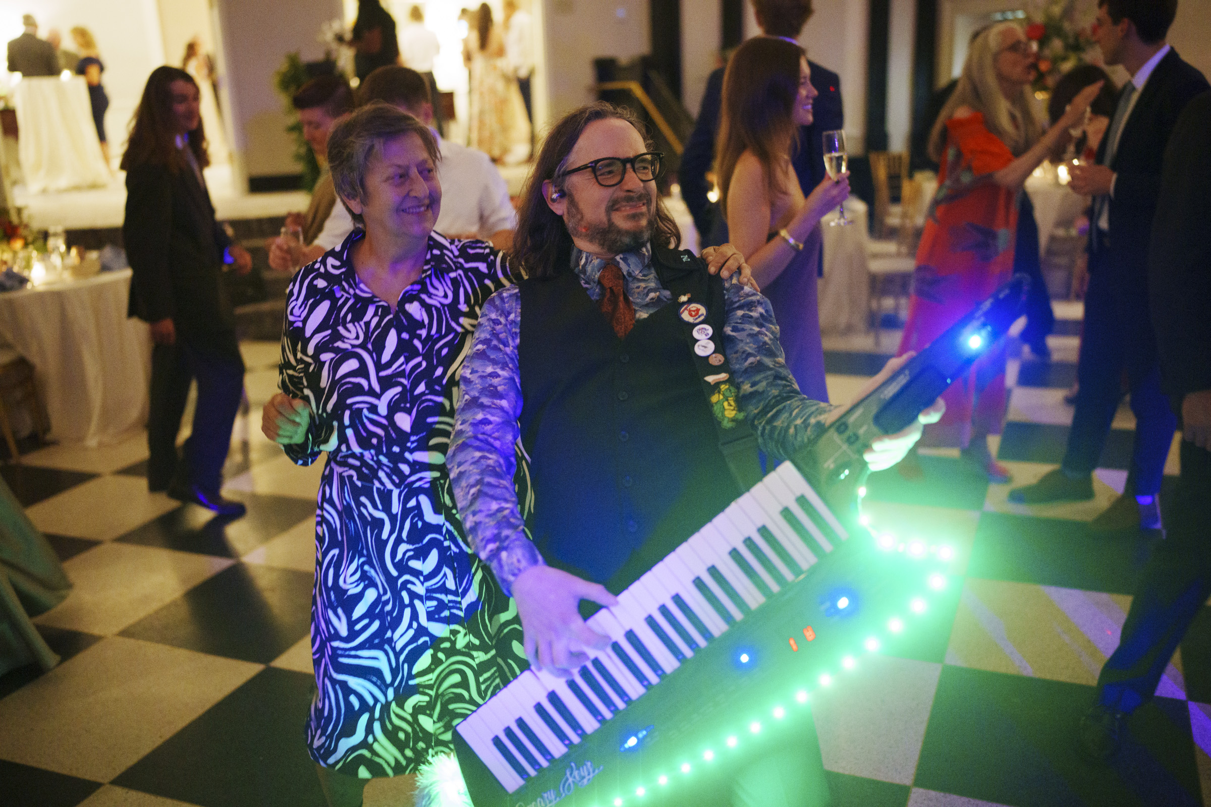 A man in a patterned shirt plays a glowing keytar while smiling, as a woman in a black and white dress dances beside him at a lively indoor party during an Elegant Spring Celebration at the Carolina Inn Wedding in Chapel Hill. By award-winning Raleigh wedding photographer, Joe Payne.